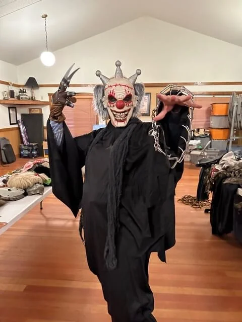 Person in a clown costume with a spooky mask, crown, and red nose, holding a chain in one hand and a prop tool in the other, standing in a room with tables and kitchen shelves.