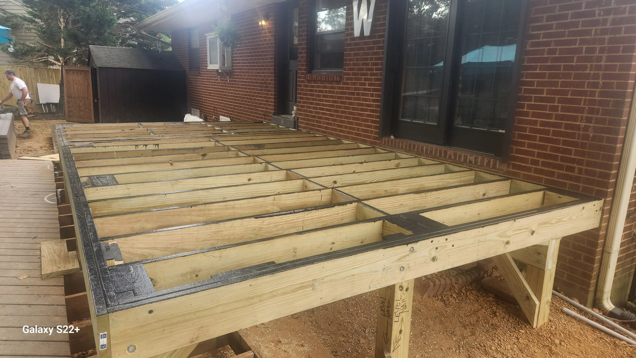 A construction site showing the wooden framing for a deck attached to a brick house with a door and windows. A man is working in the background.