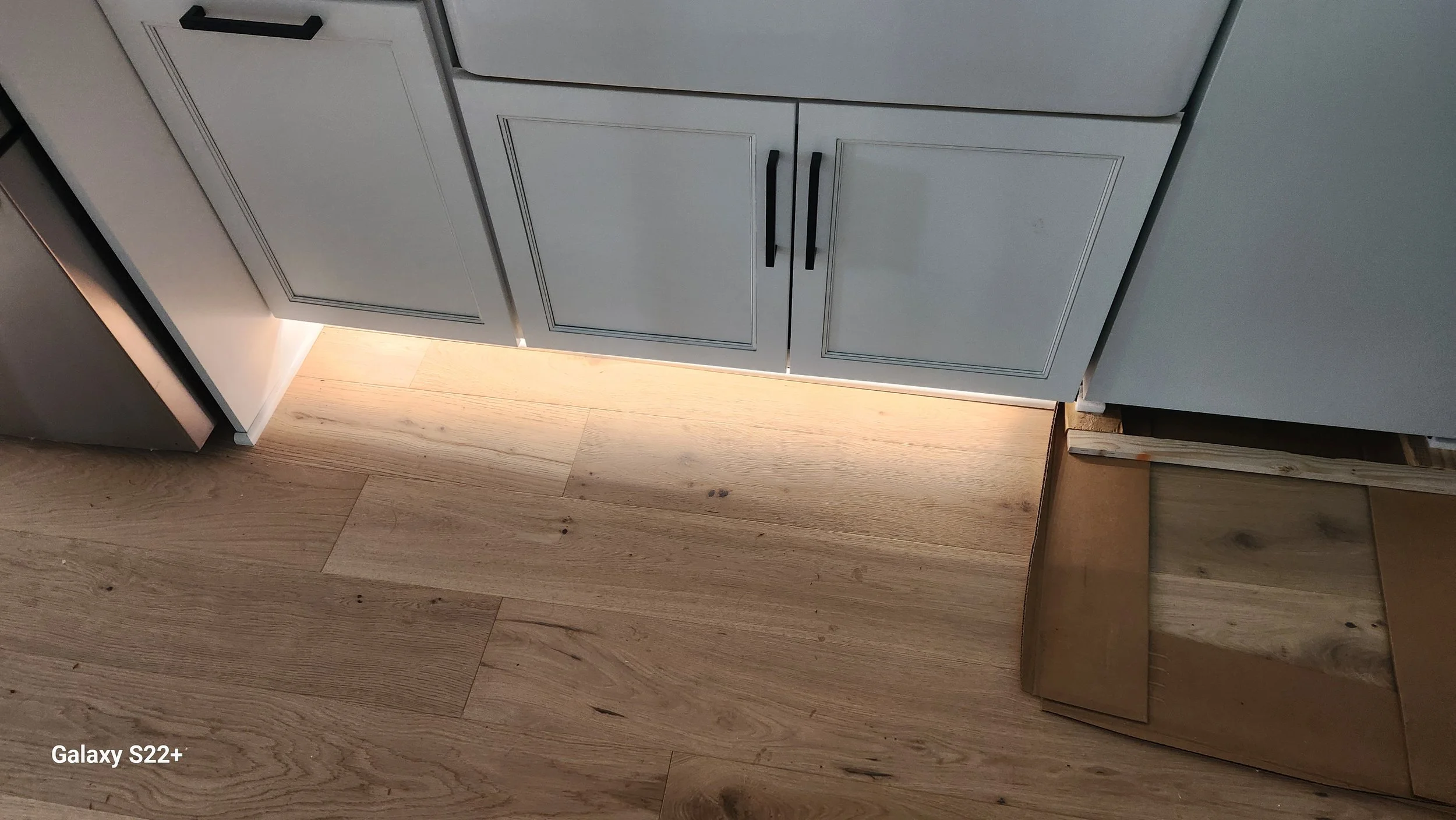 View of kitchen cabinets with black handles, light-colored wooden floor, and illuminated baseboard lighting beneath the cabinets. Part of a cardboard box is visible on the right side.