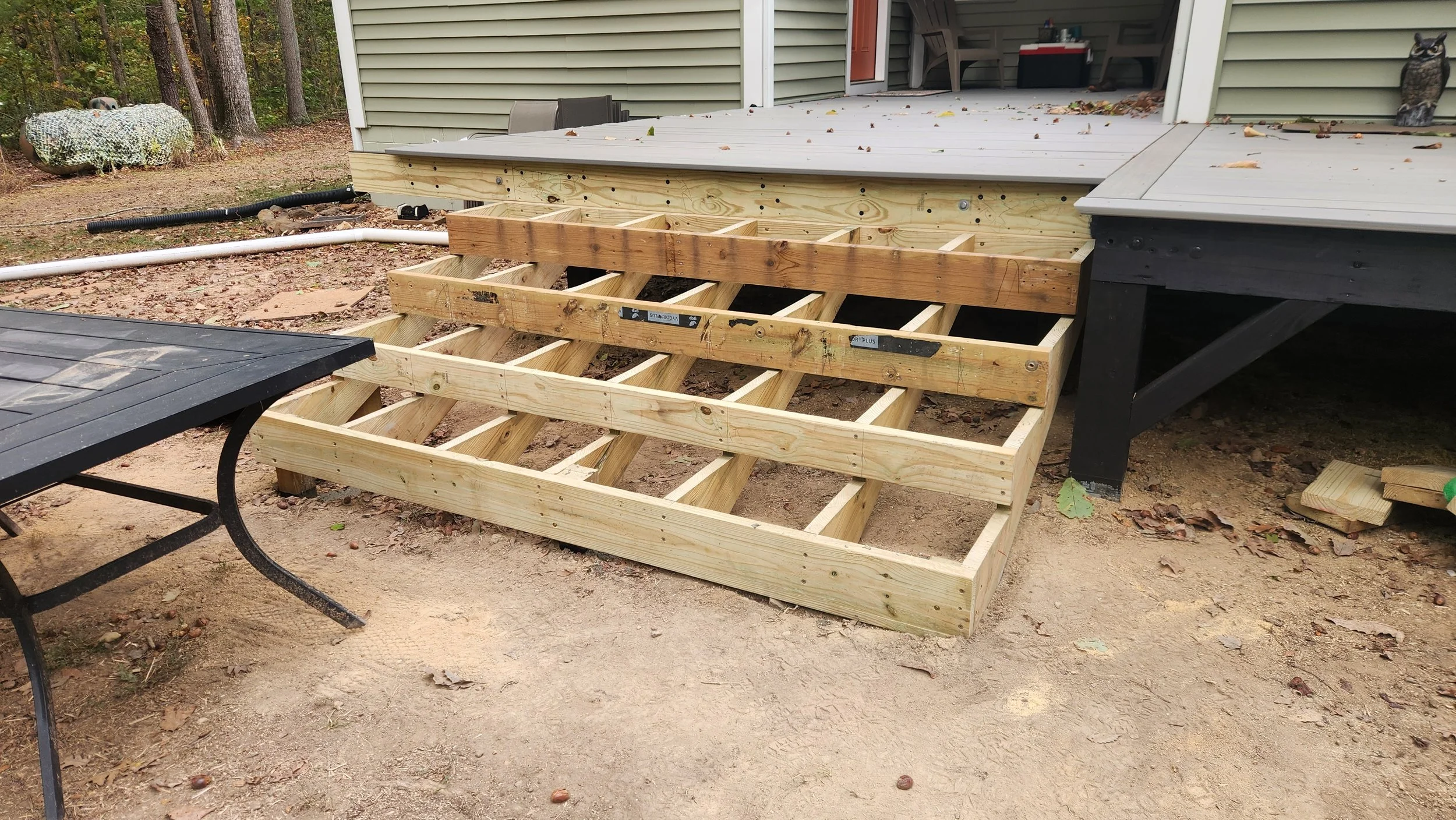 Wooden deck stairs under construction next to a house, with a black outdoor table in the foreground and trees in the background.