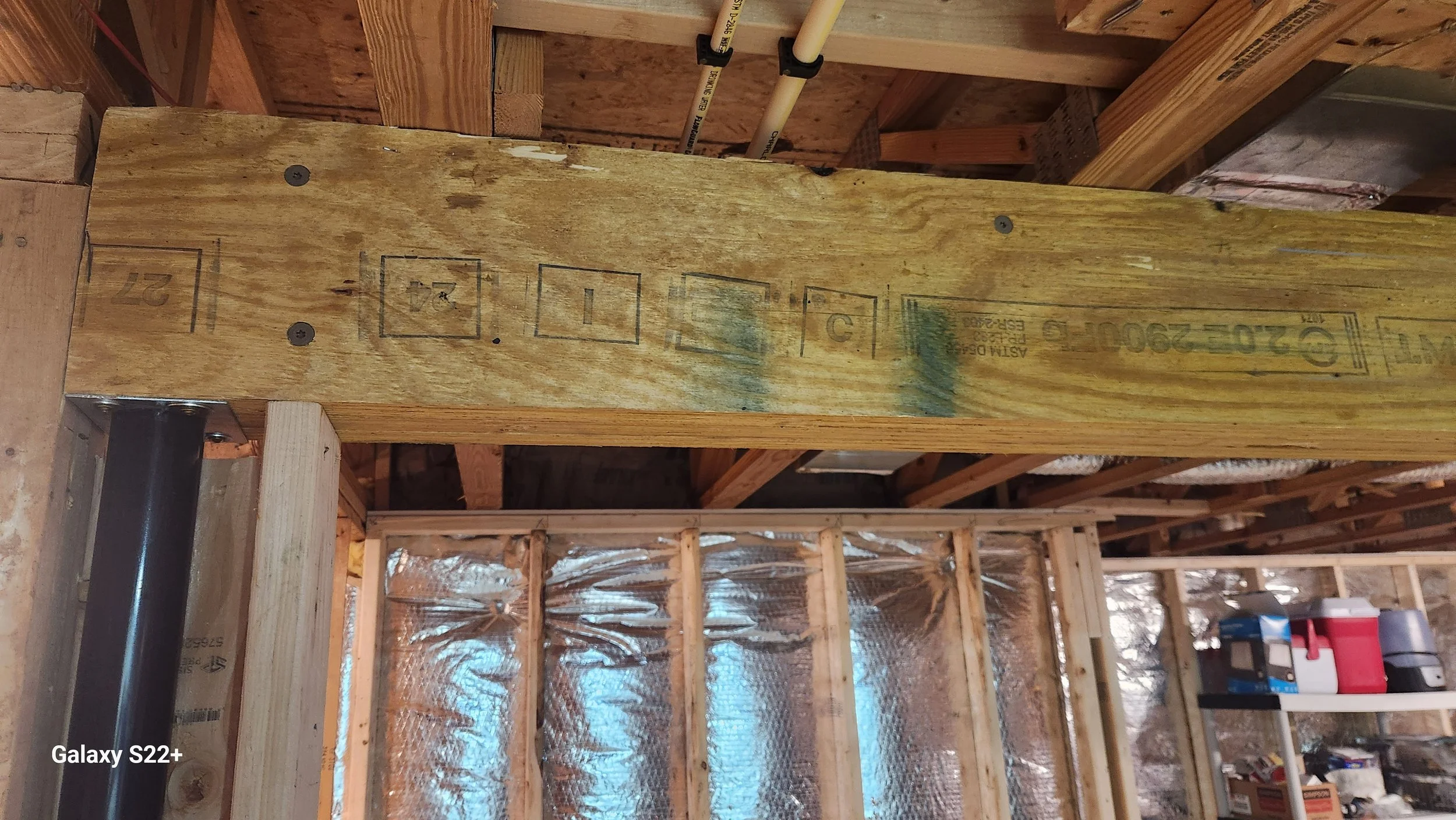 Wooden framing in a construction site with a large wooden beam marked with power and size specifications, and a spray of green paint. Insulation and construction tools are visible in the background.