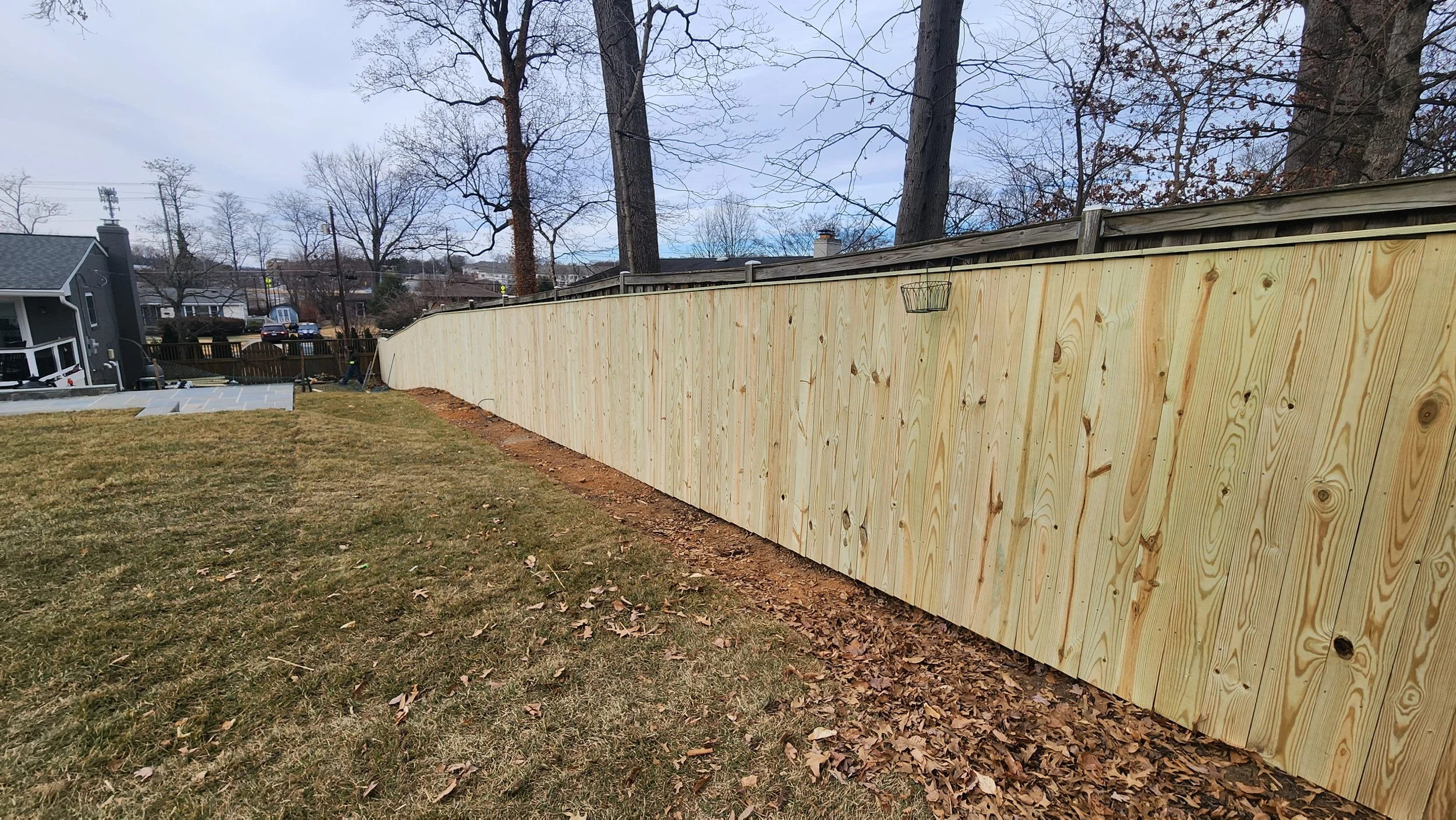 A newly installed wooden privacy fence along a backyard, with a grassy lawn and some fallen leaves in the foreground, and neighboring houses and trees in the background.