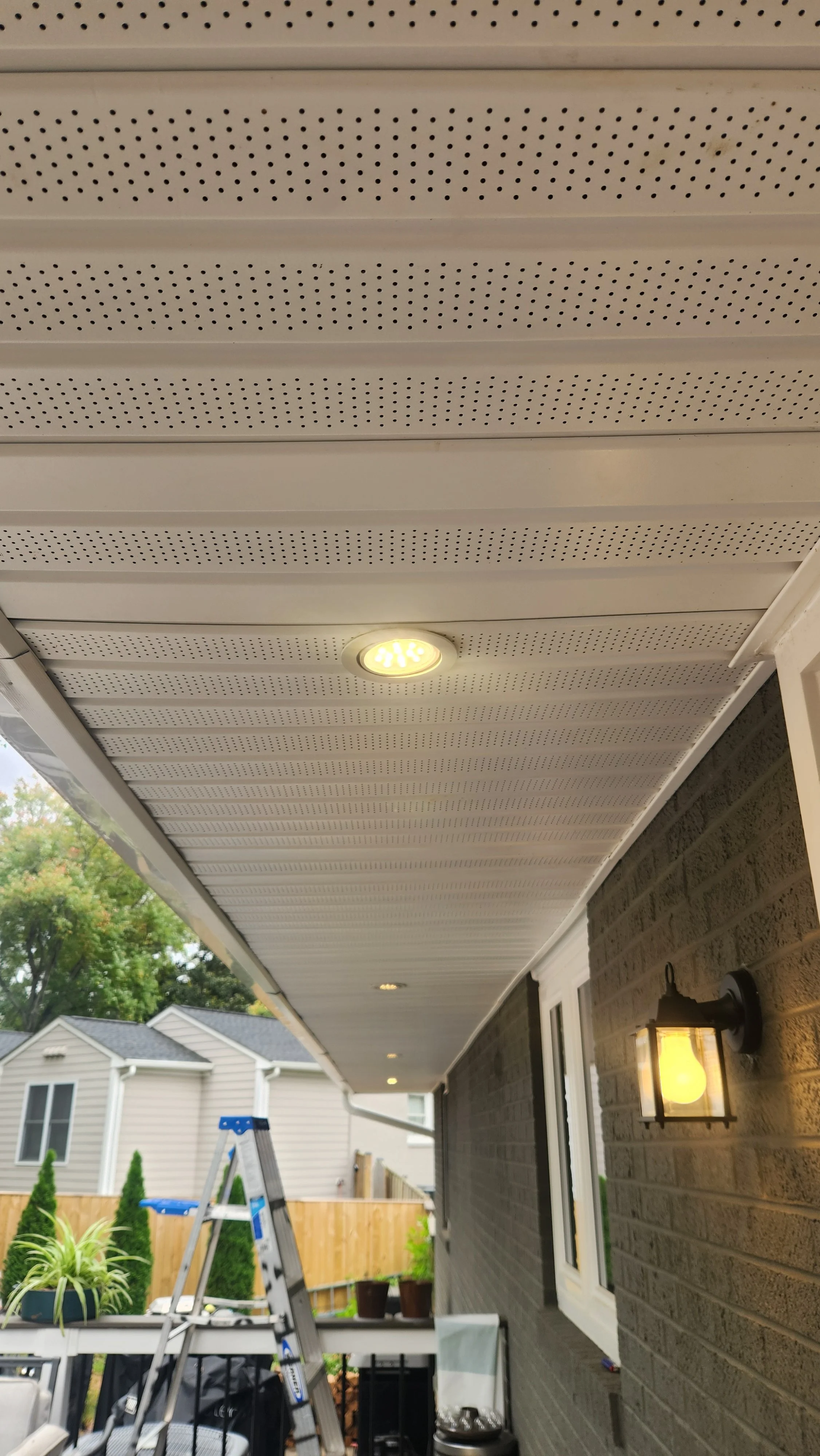 View of a house patio with a white perforated soffit ceiling, installed recessed lighting, exterior wall with a wall-mounted lantern, patio furniture, ladder, and potted plants.