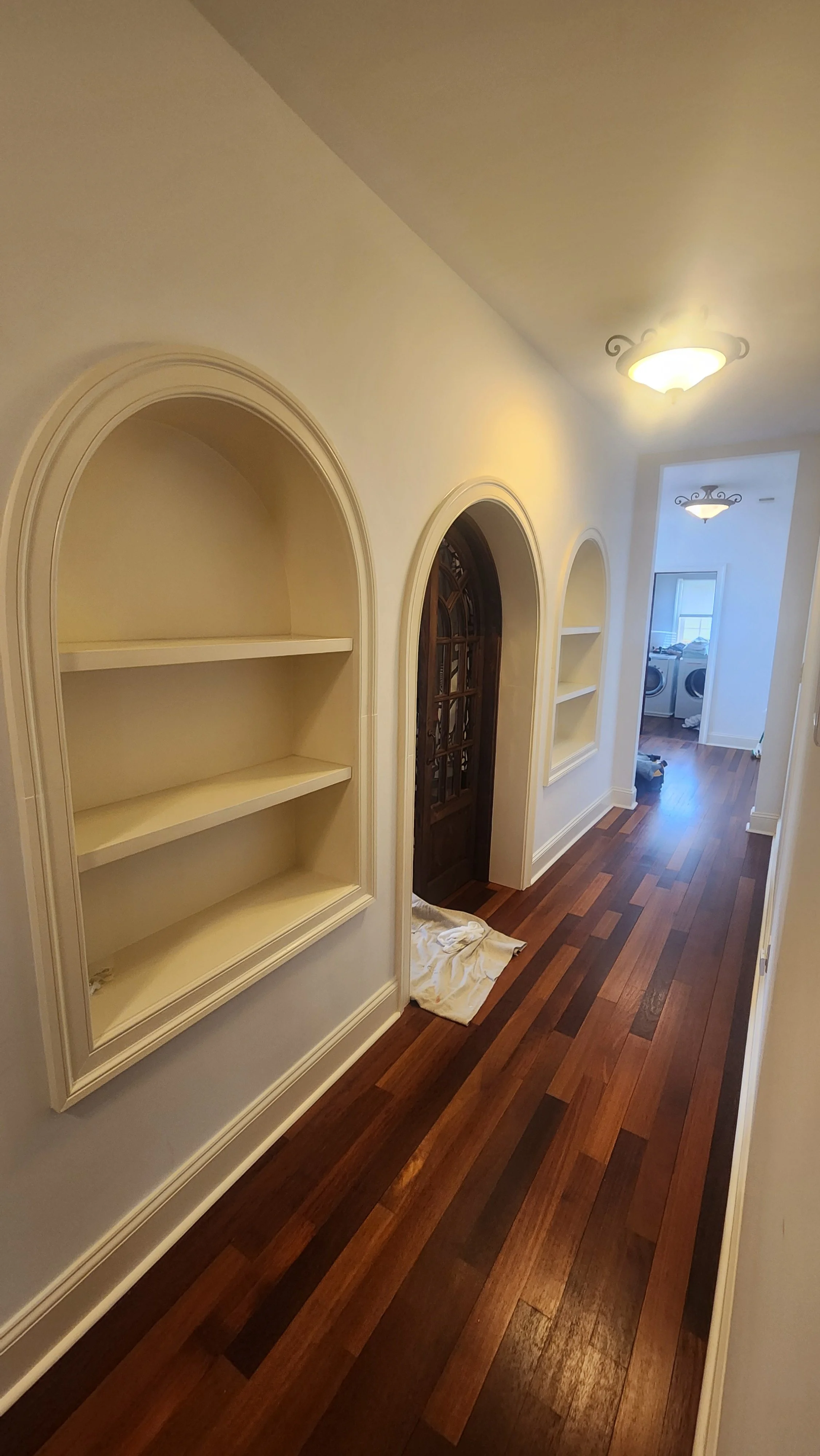 Hallway with built-in wall shelves, hardwood flooring, and ceiling lights, leading to a laundry room with washers and dryers in the background.