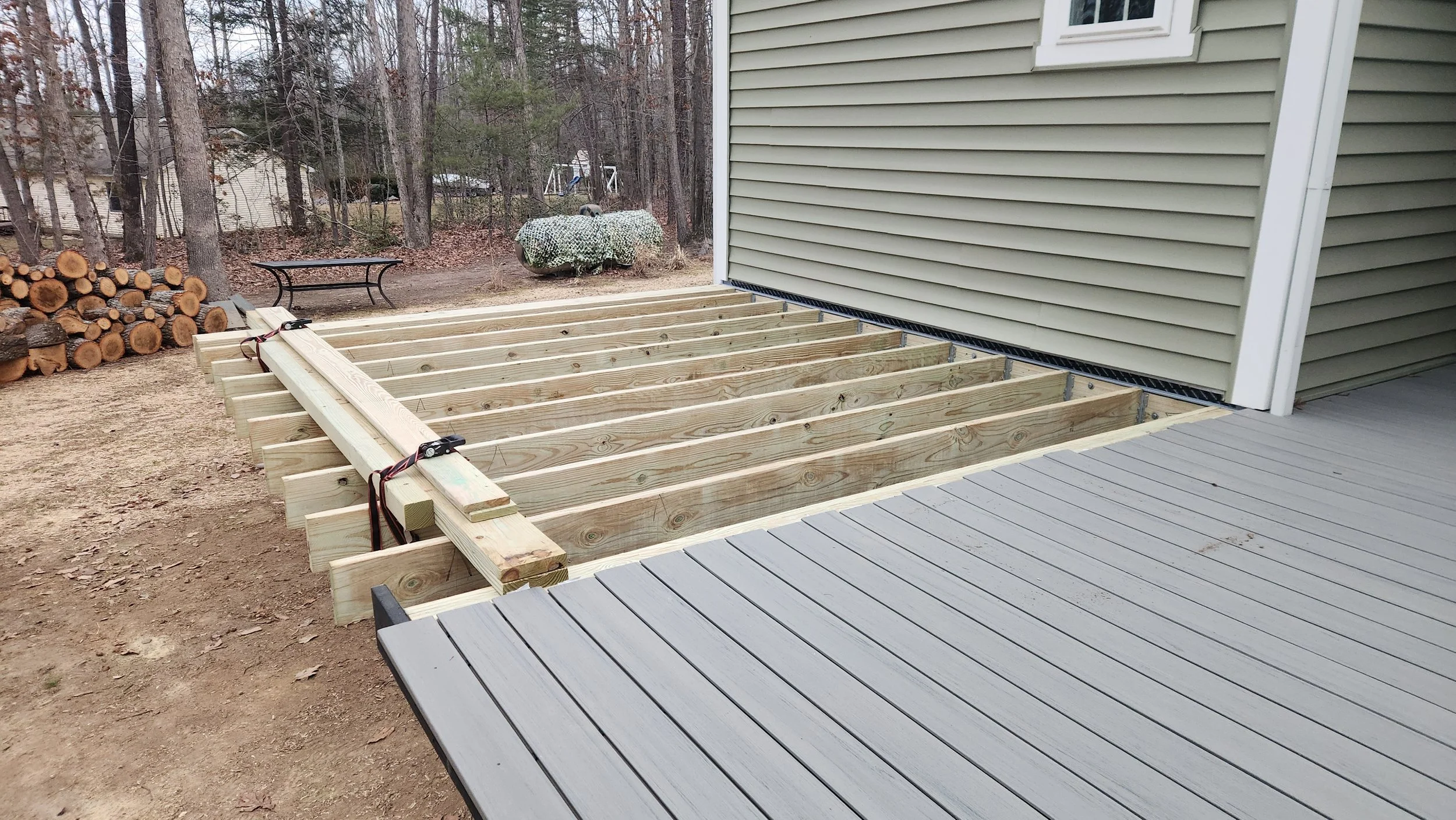 Newly constructed deck with wooden framing, attached to a house, with a gray composite deck surface nearby, in a backyard with trees and logs.