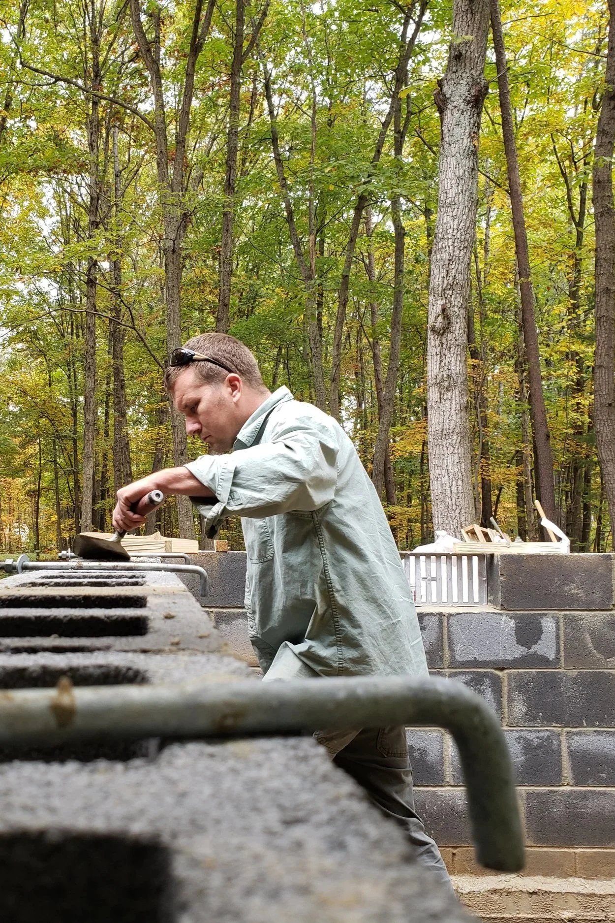 A man working on a brick outdoor grill in a forested area during daytime.
