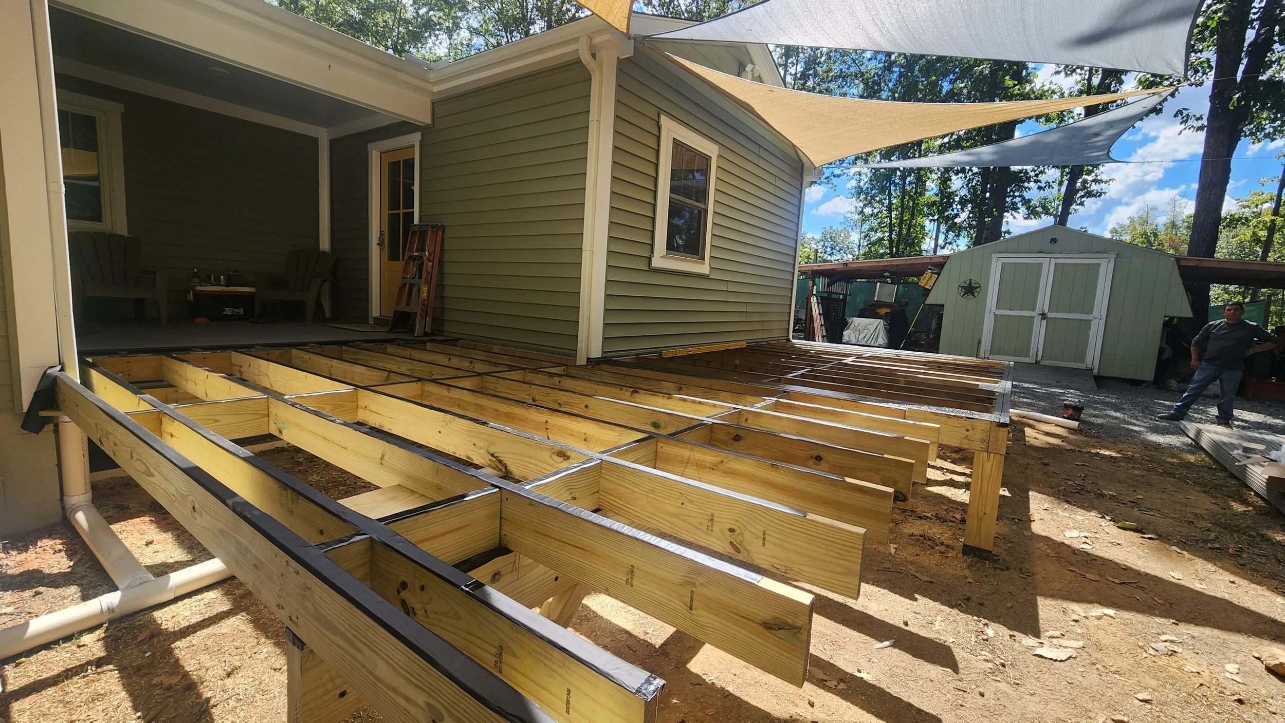 A residential backyard under construction, featuring a wooden deck framework with support beams and joists, adjacent to a house with a porch, and a green shed in the background, with a man standing nearby.