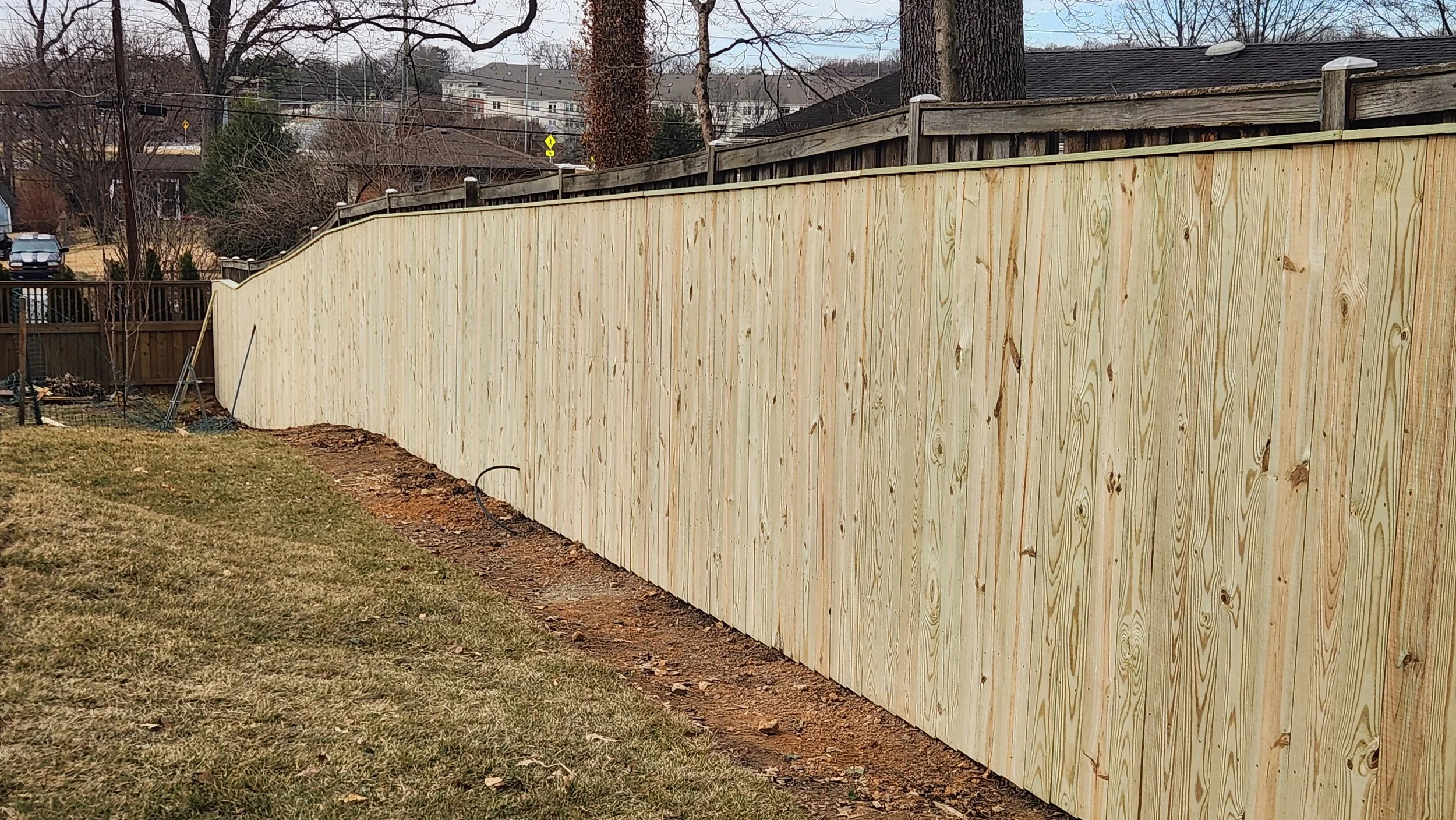 A new wooden privacy fence installed in a backyard, with some trees and houses visible beyond it.