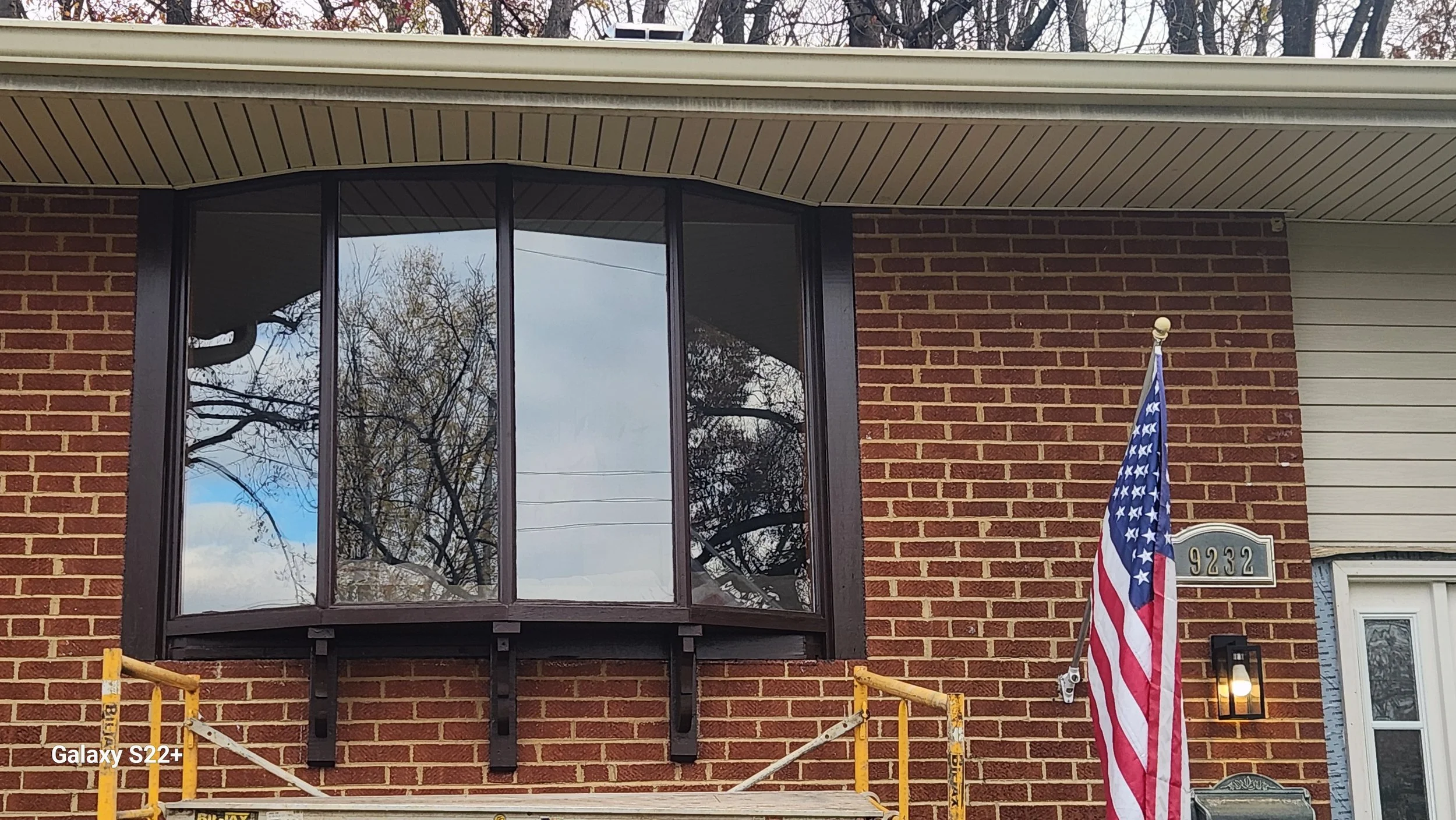 Brick house with a large bay window reflecting trees, American flag on a pole, house number 9232, and a lit exterior lantern.