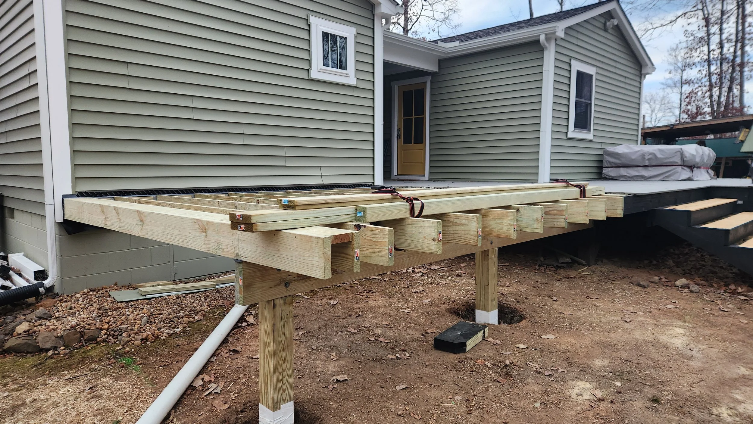 A wooden deck under construction attached to a light-colored house. The deck's framing is visible, with some boards stacked nearby and ready for installation. The ground beneath is bare dirt, and stairs lead down from the deck to the yard.