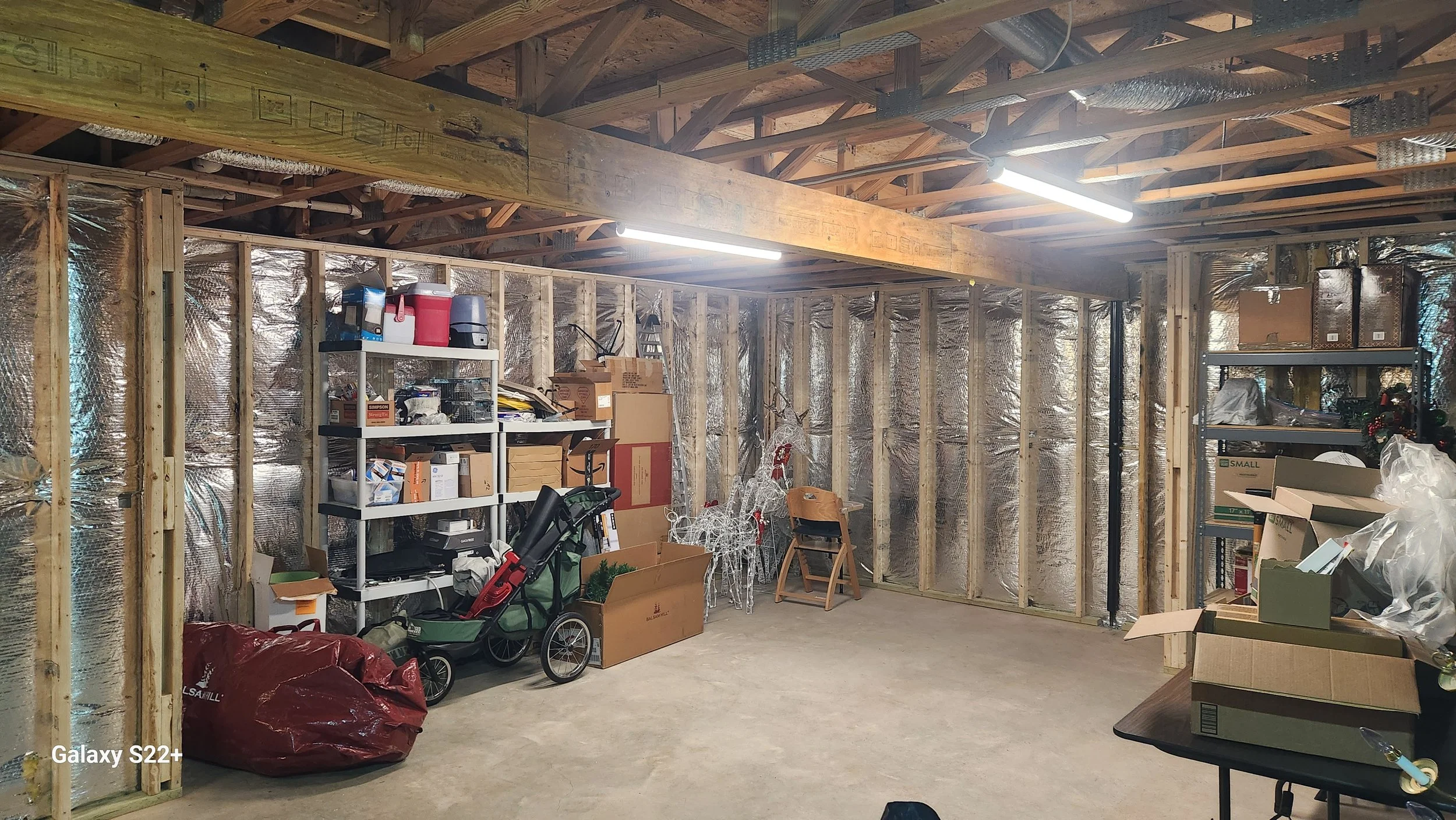 An unfinished garage with exposed wooden beams and foil insulation on the walls. It contains shelves filled with boxes, containers, and miscellaneous items, along with a folded stroller, cardboard boxes, and Christmas decorations.