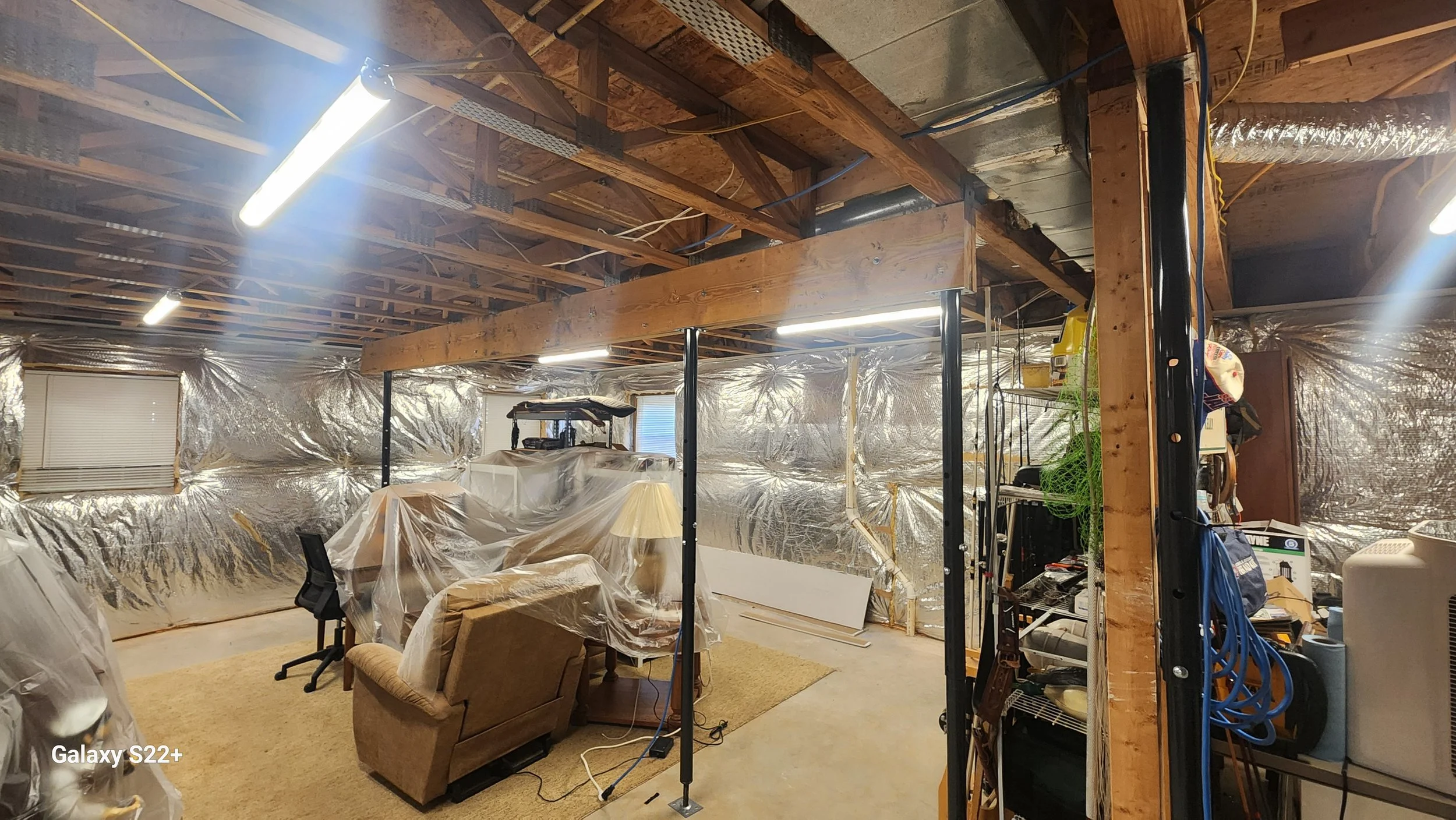 A cluttered basement with unfinished wooden ceiling beams, covered ductwork and drywall, and reflective insulation on the walls. There are various pieces of furniture including a sofa, a chair, and a lamp, all partly covered with plastic. The space h