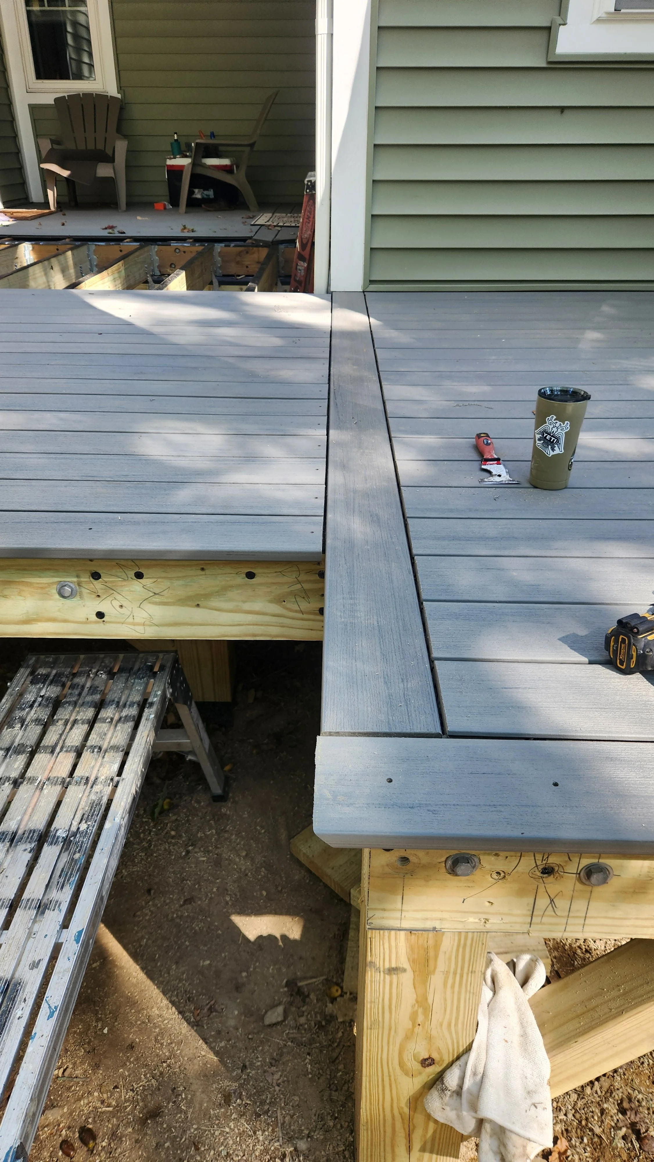 Construction of a wooden deck with gray composite planks, partially built, with tools and materials on top, and a partially completed section revealing the underlying wooden structure.