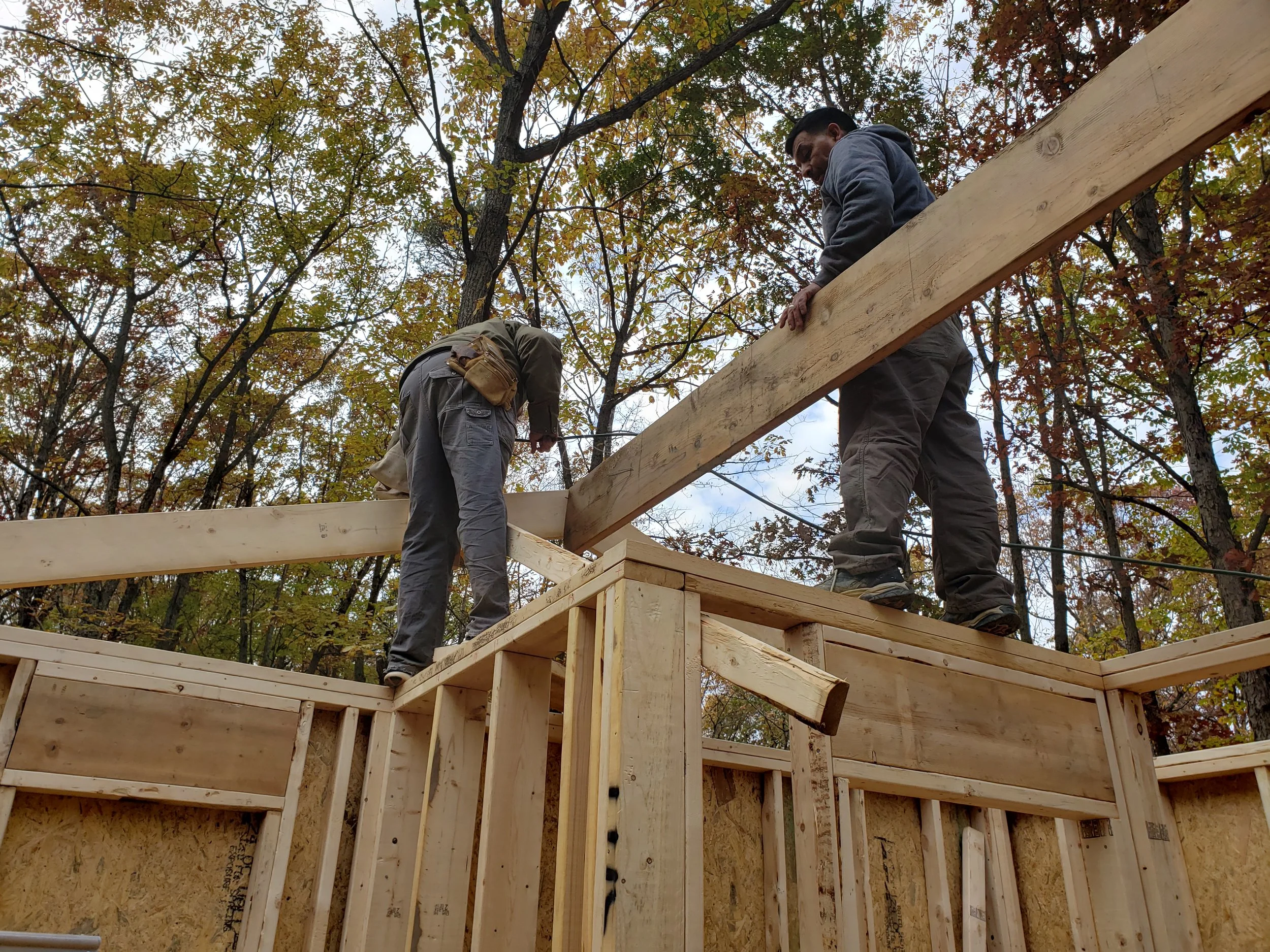 Two construction workers are working on a wooden structure in a forested area, positioning and securing large beams for a building frame.