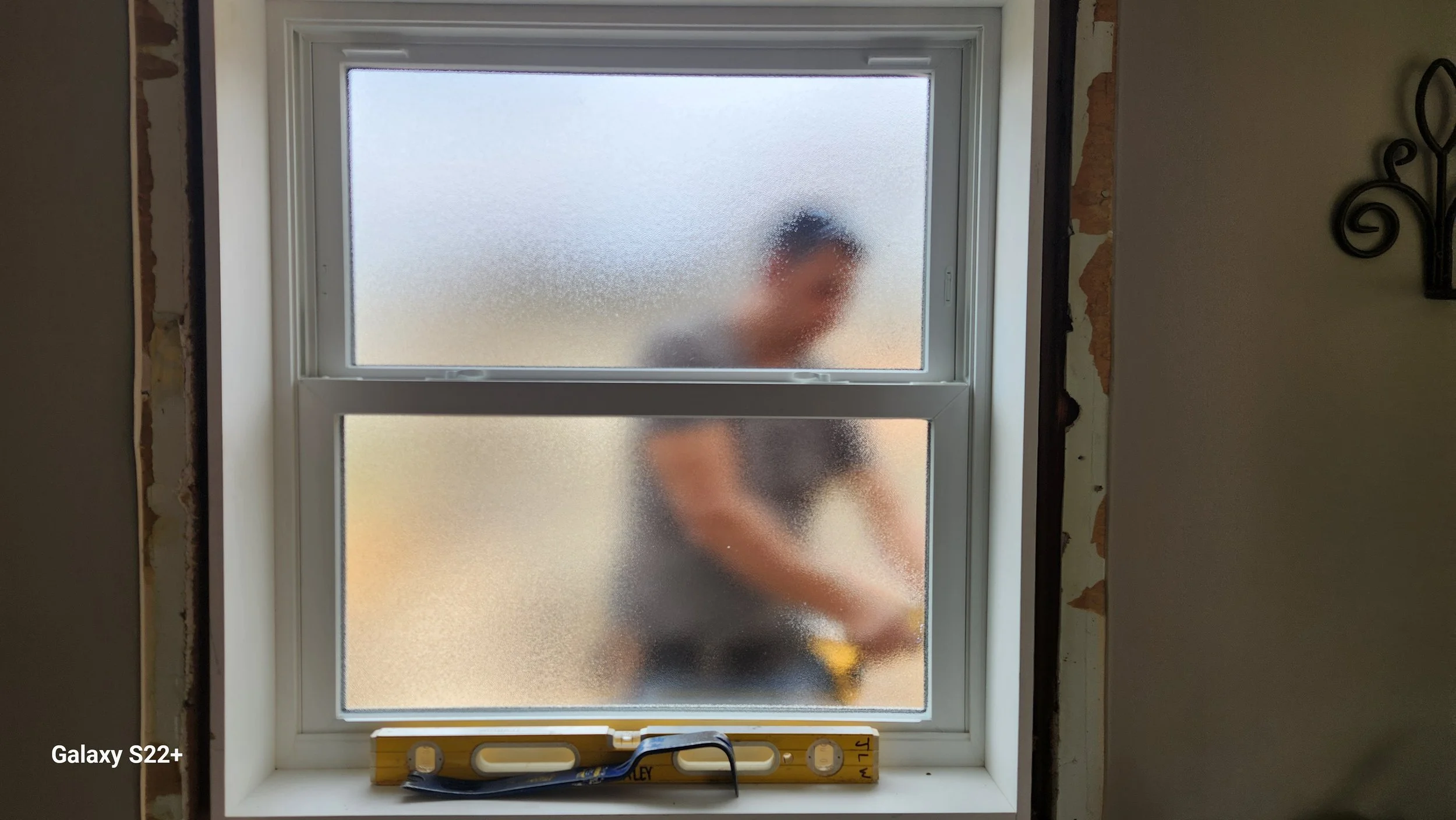 A blurry person is seen through a frosted window, standing outside and working on a house. The inside wall beside the window shows exposed drywall edges, and there is a level and a paint scraper on the white window sill.