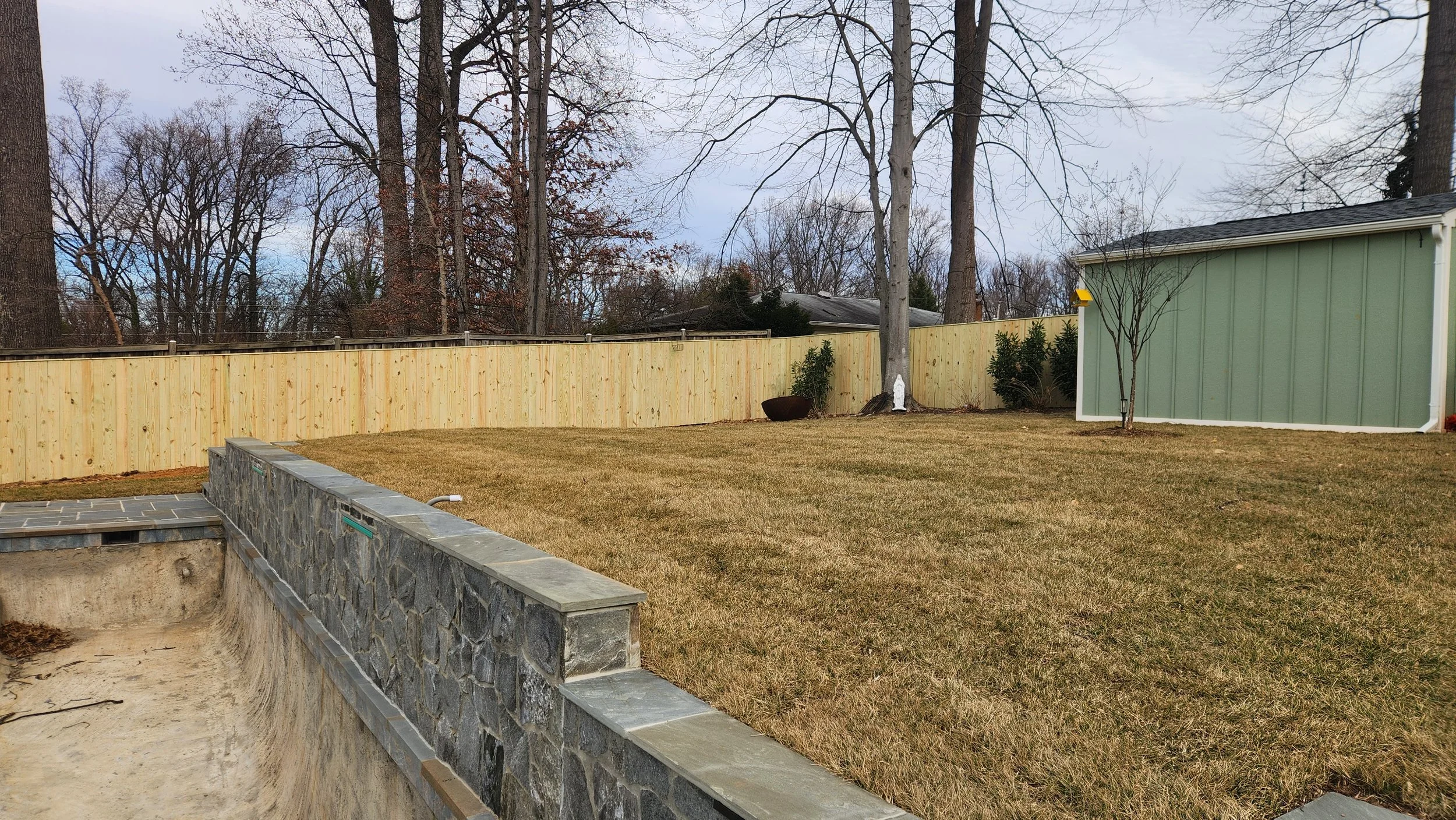 Backyard with a grass lawn, stone-bordered in-ground pool under construction, wooden fencing, small trees, and a green shed.