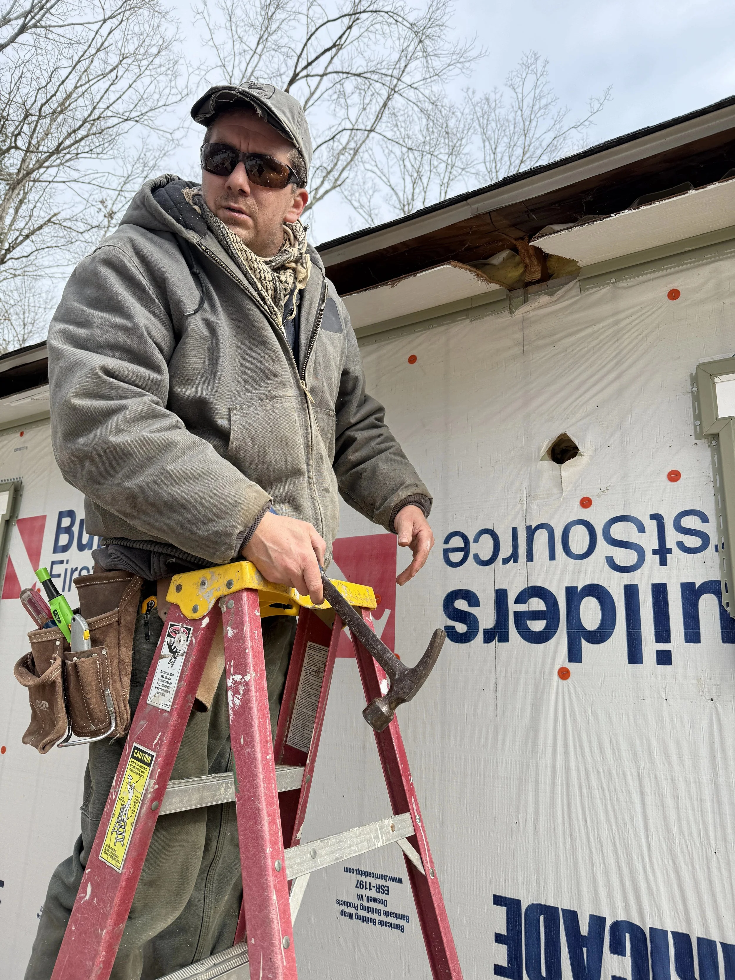 A man wearing sunglasses, a cap, and work clothing stands on a red ladder working on the exterior wall of a building under construction or renovation.