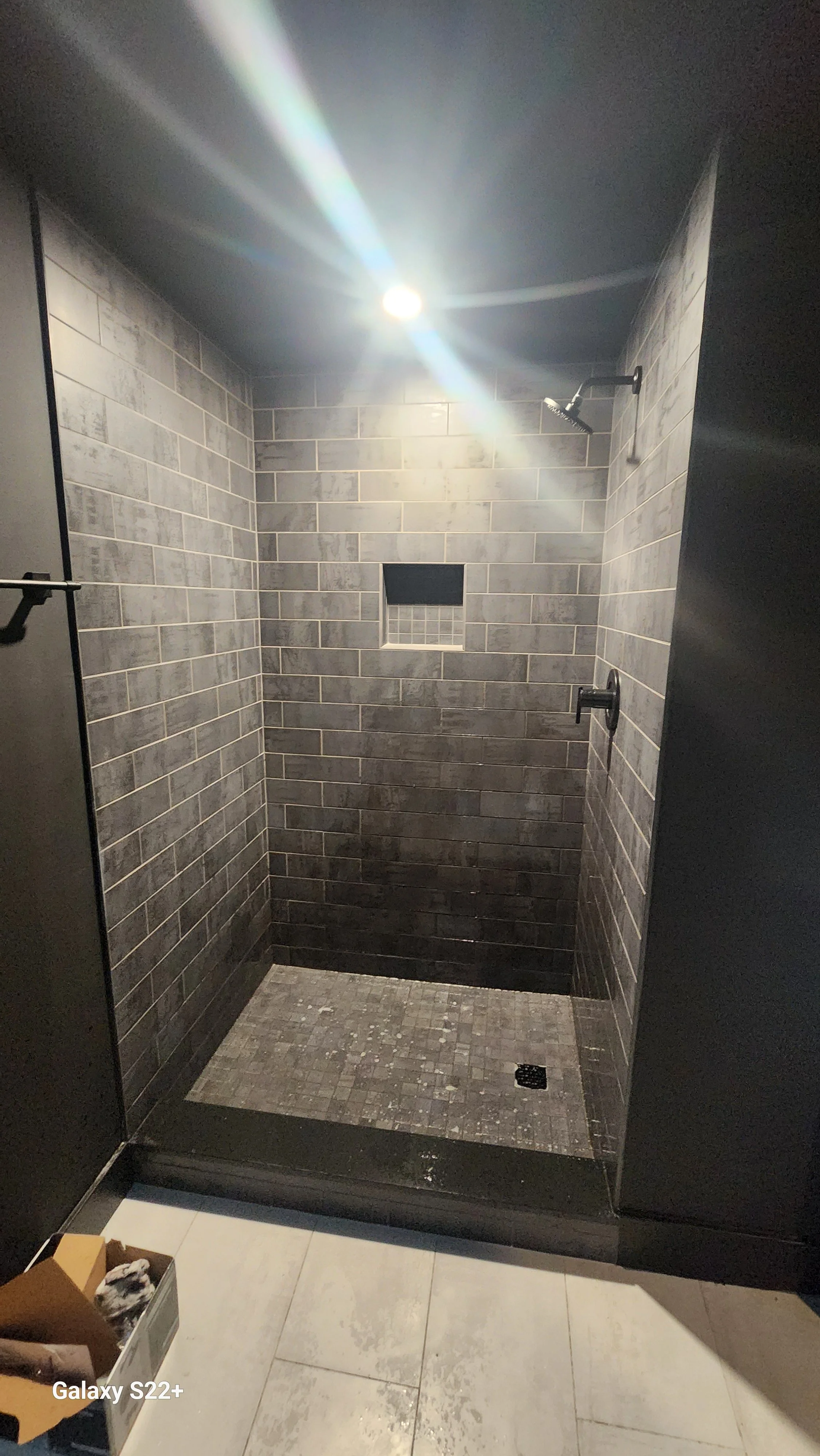 A modern walk-in shower with dark gray brick tiles on the walls, a small built-in tile shelf, a ceiling showerhead, and a floor drain, with a concrete-colored tiled floor outside.