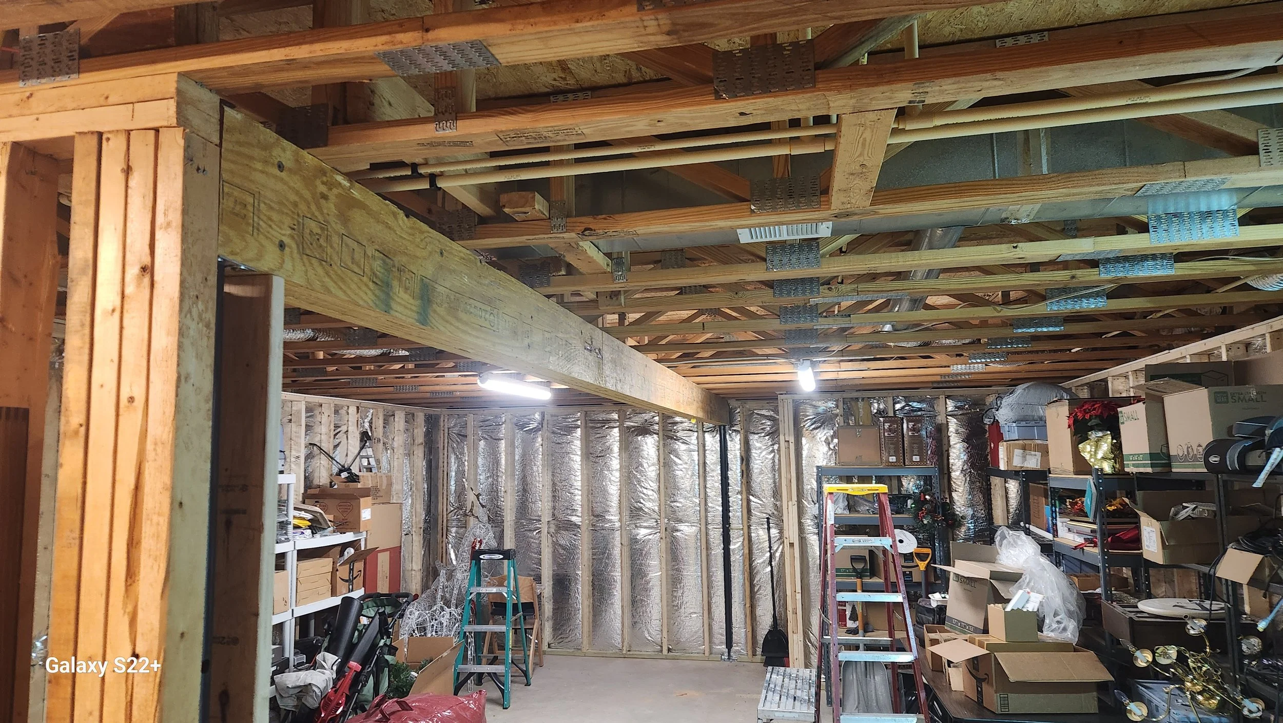 Interior of a garage under construction with exposed wooden framing, shelves with boxes and tools, a ladder, and construction materials.