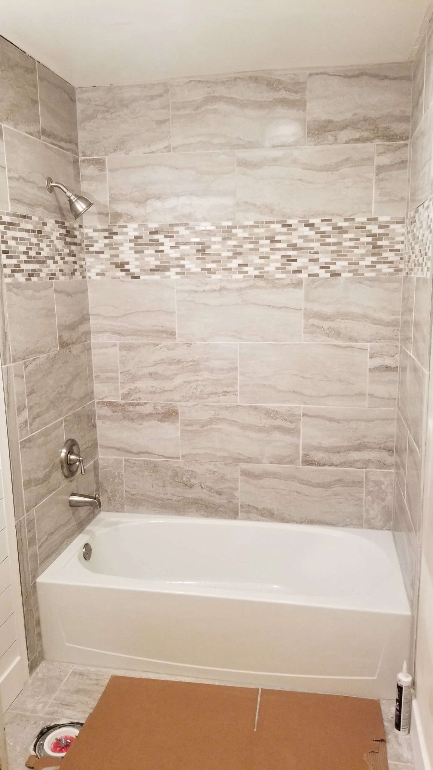 Bathroom shower with beige and gray stone tiles, a mosaic accent strip, a bathtub, and a bottle of cleaning product on the floor.