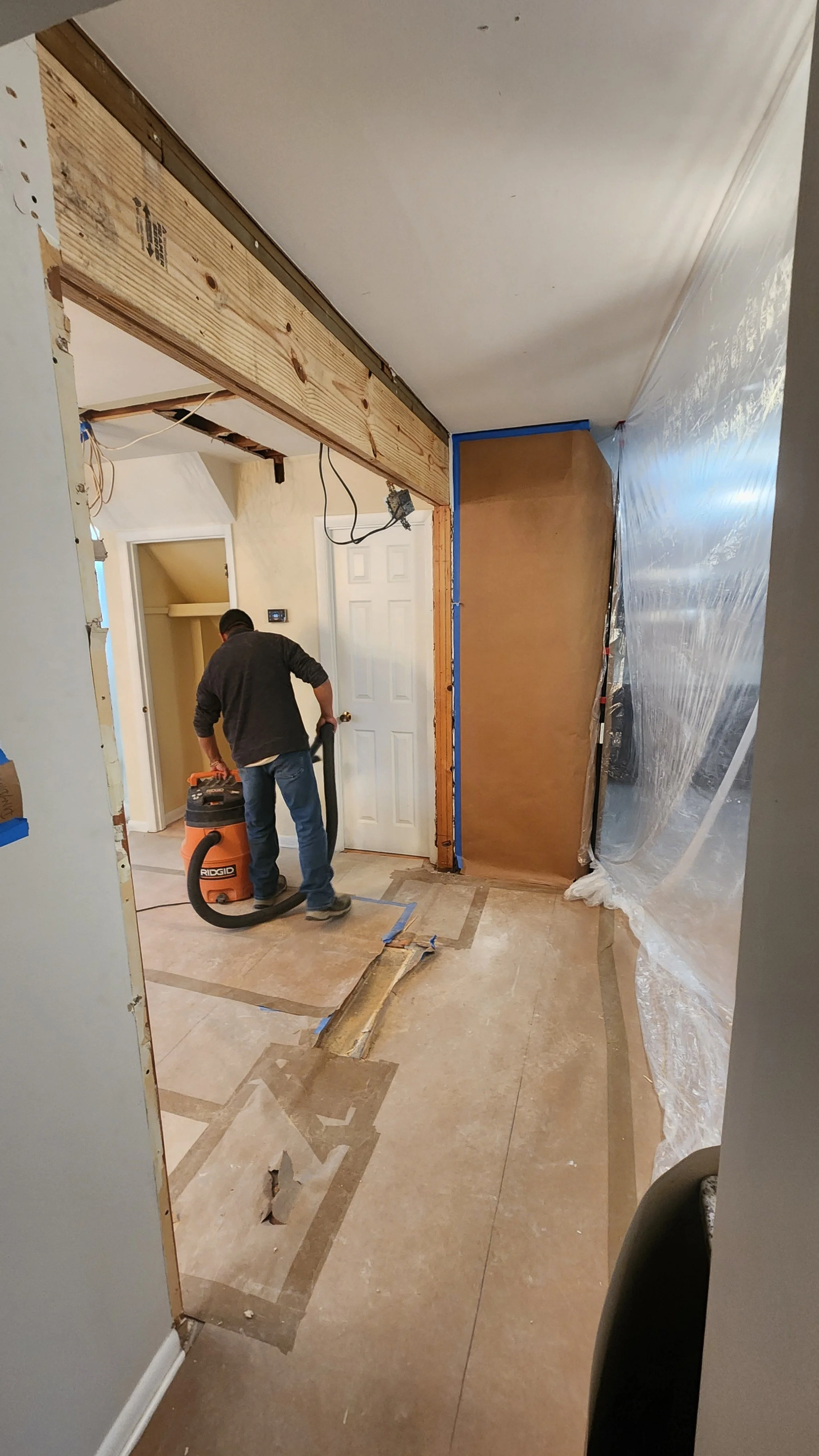 Construction worker vacuuming debris in a room undergoing renovation, with exposed framing and partially removed flooring.