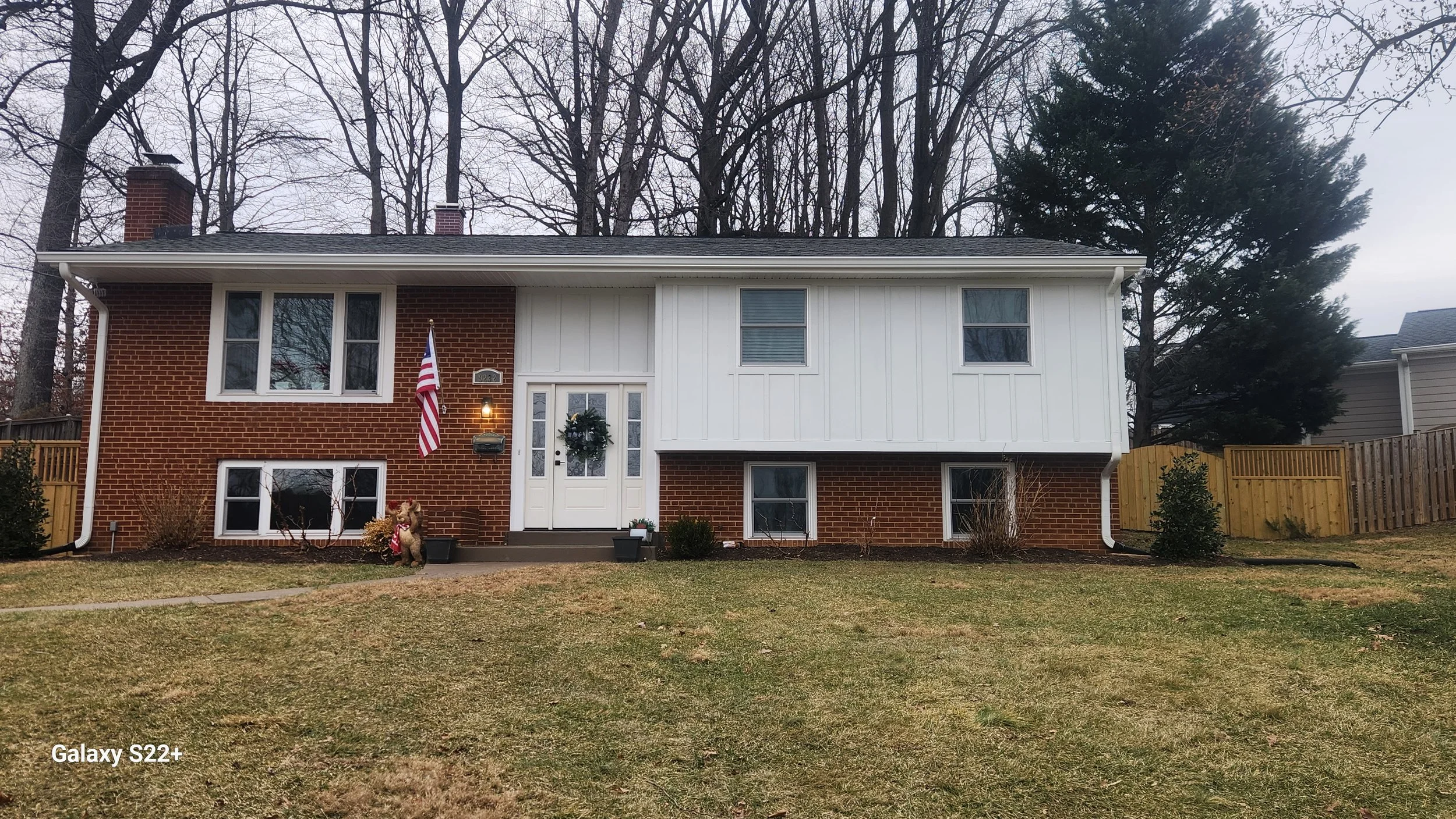 Front view of a two-story house with a brick exterior on the lower level and white siding on the upper level. The house has multiple windows, a white front door with a wreath, an American flag near the door, and a hanging light. There are small plant