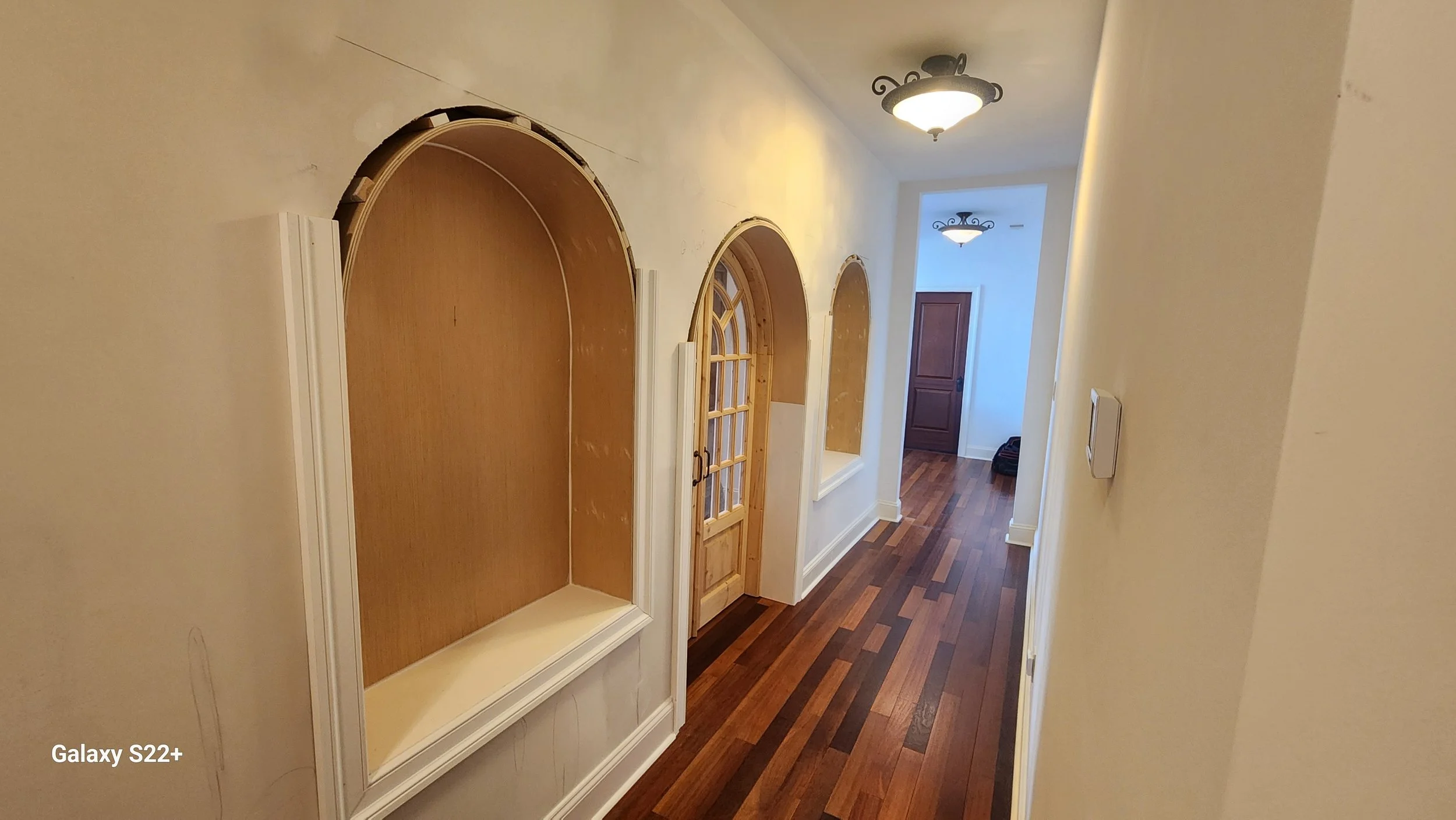 A hallway with wooden flooring, wall niches with wood backing, and a wooden door at the end. Overhead ceiling lights illuminate the corridor.