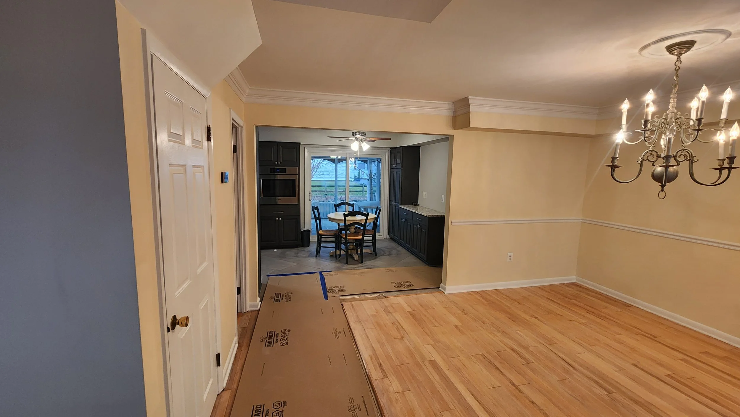 Photo of a living room with a chandelier, partially finished hardwood flooring, and a view into a kitchen with dark cabinets, a dining table, and large windows.