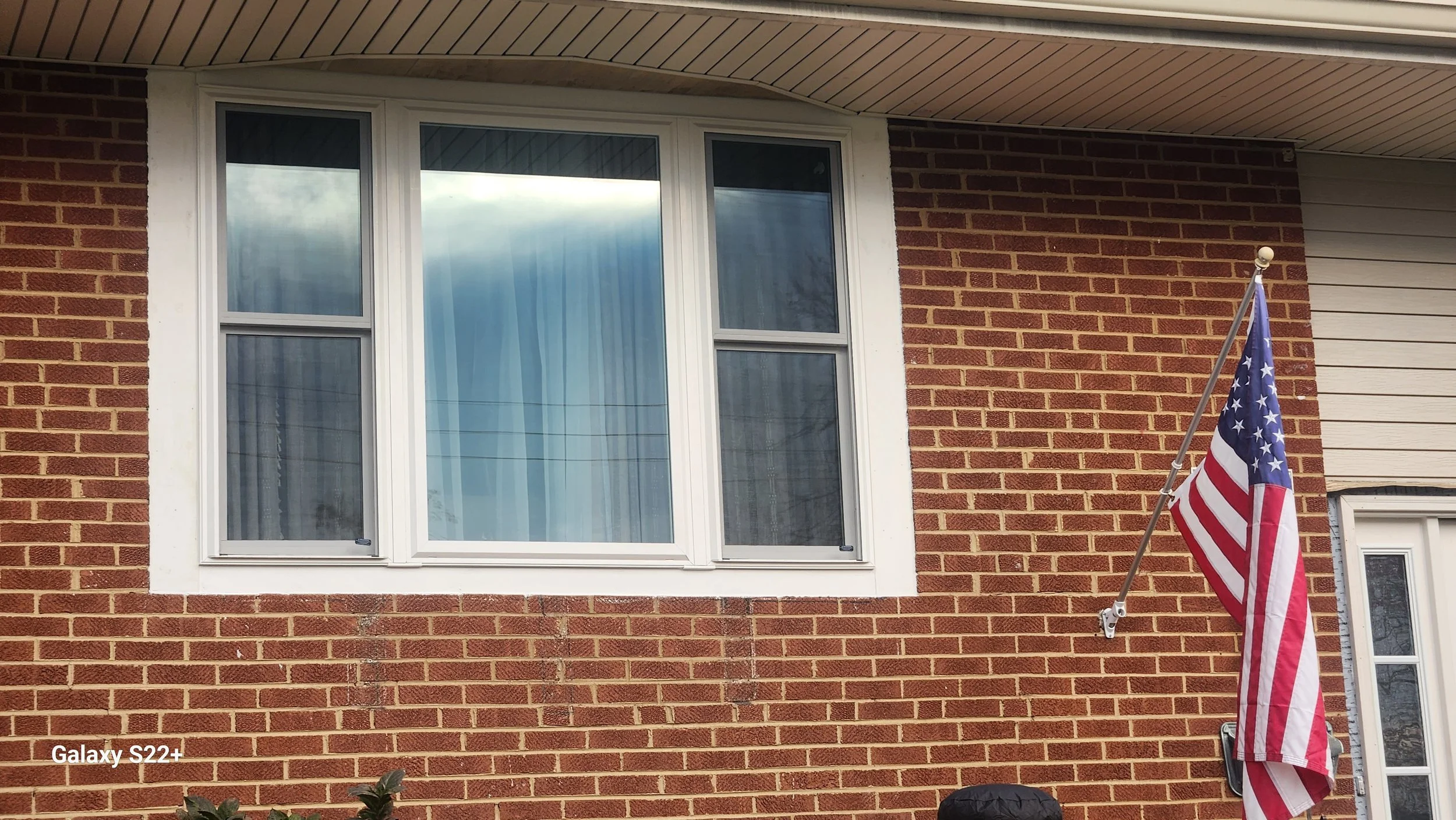 A brick house with a large window and an American flag mounted on the wall to the right of the window.