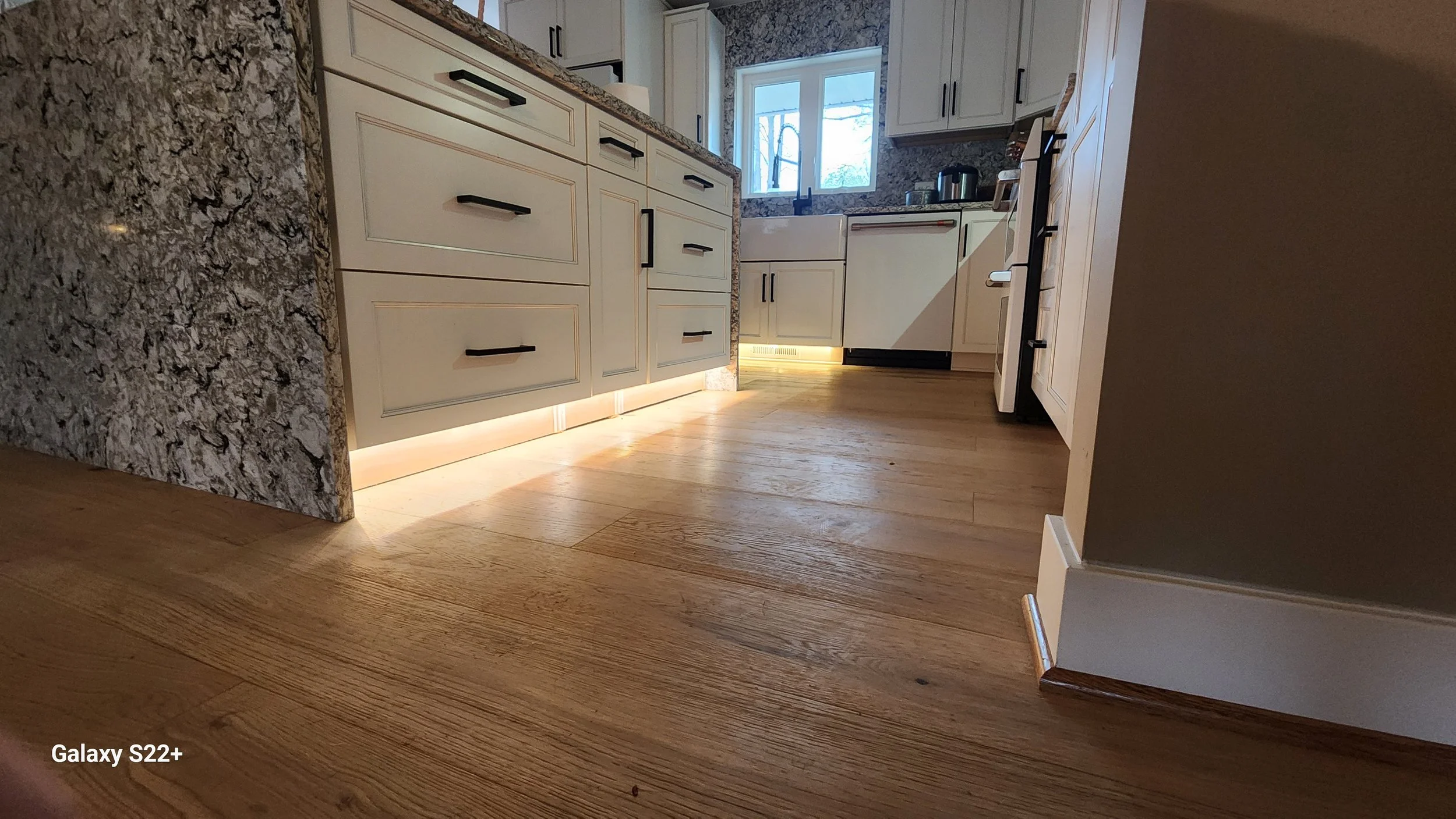 A modern kitchen with white cabinets, granite countertops, and wooden flooring. The cabinetry features black handles, and there is under-cabinet lighting illuminating the floor. A window is visible above the sink, with a few items on the counter near