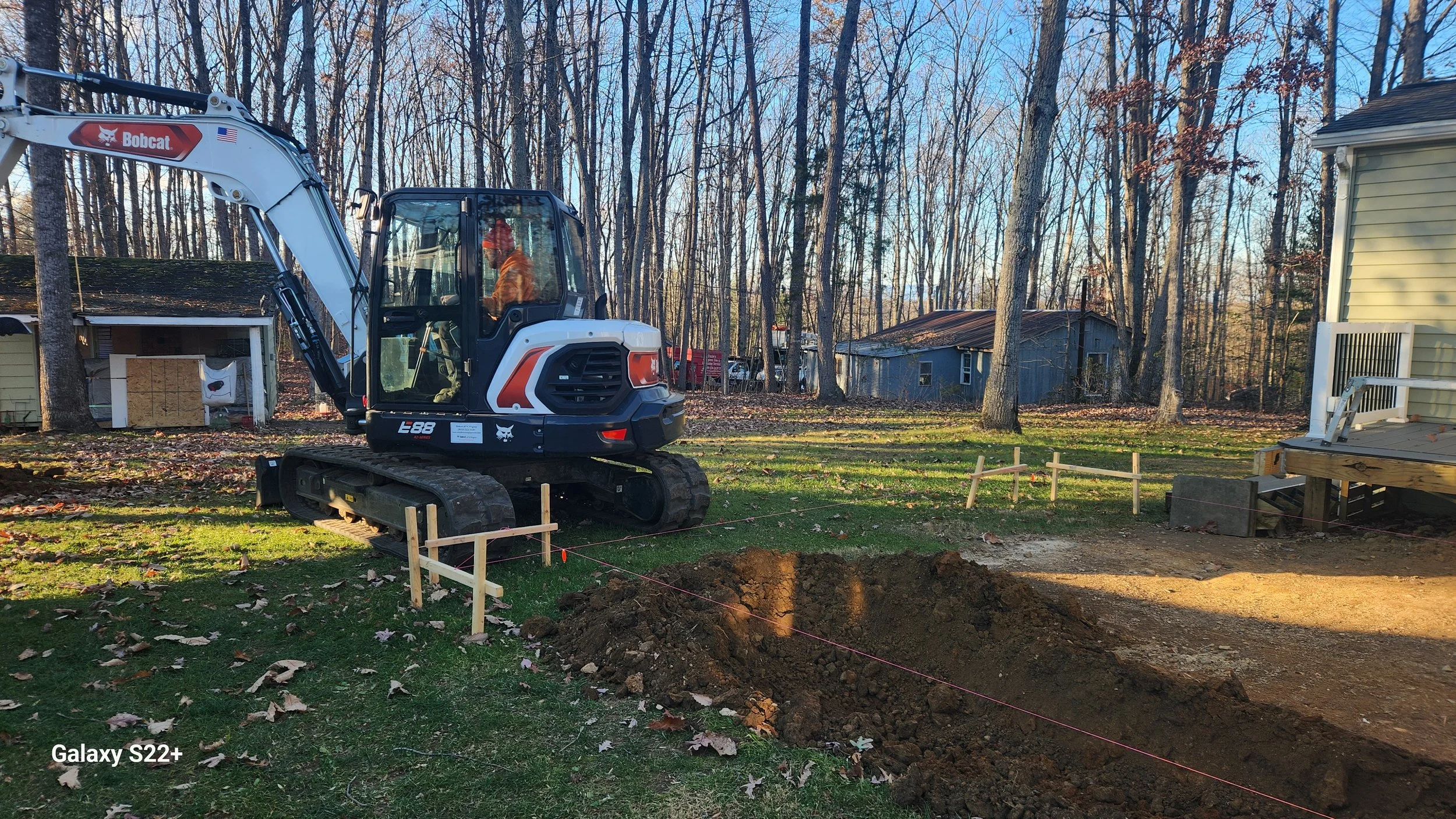 A construction site in a backyard with a small excavator digging a trench. The excavator is white with orange and black accents, and the operator is inside. The area is marked off with small wooden stakes and pink string. The background features tree