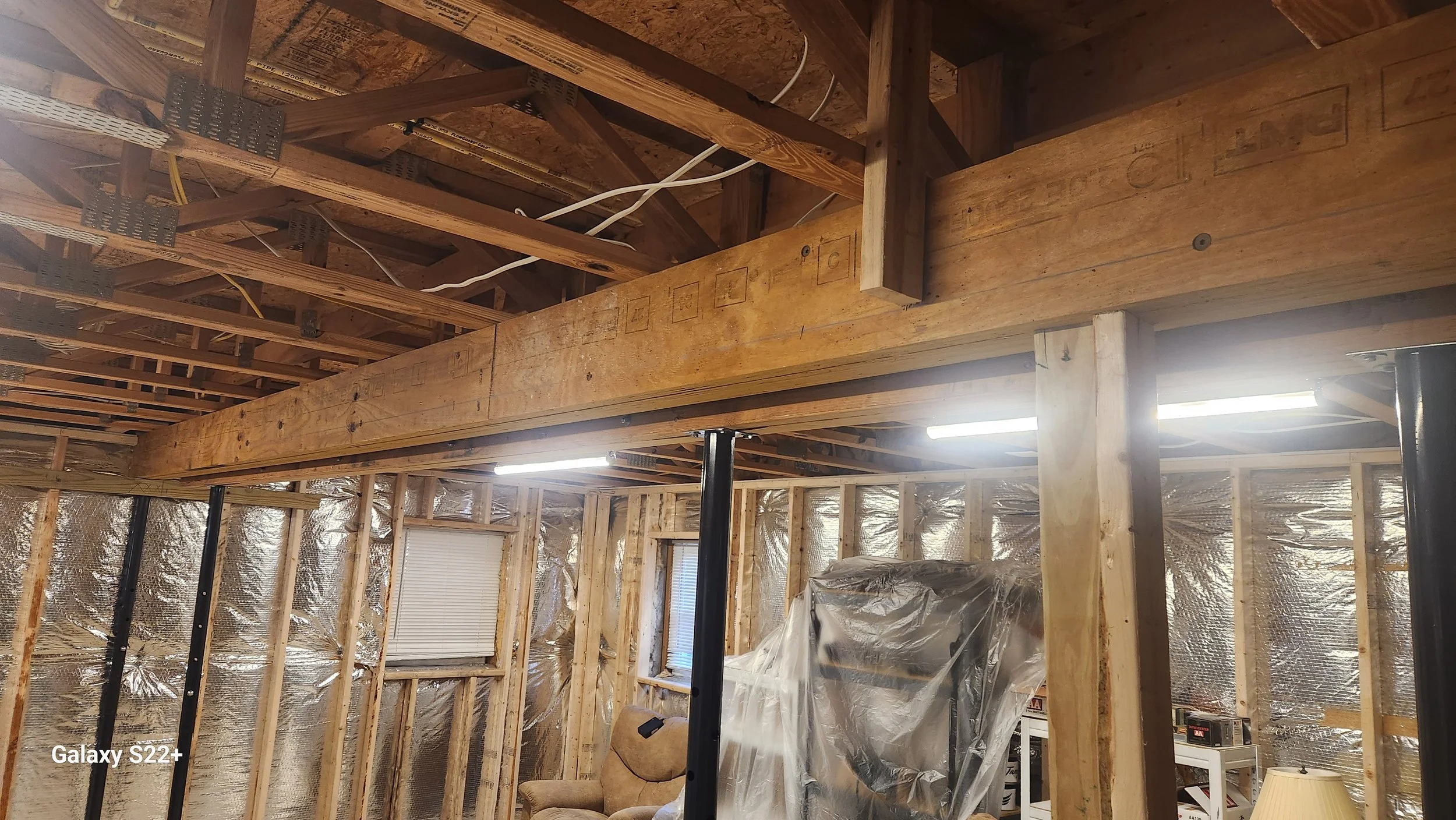 Interior of a house under construction with exposed wooden framing, insulation, and a black support pole.