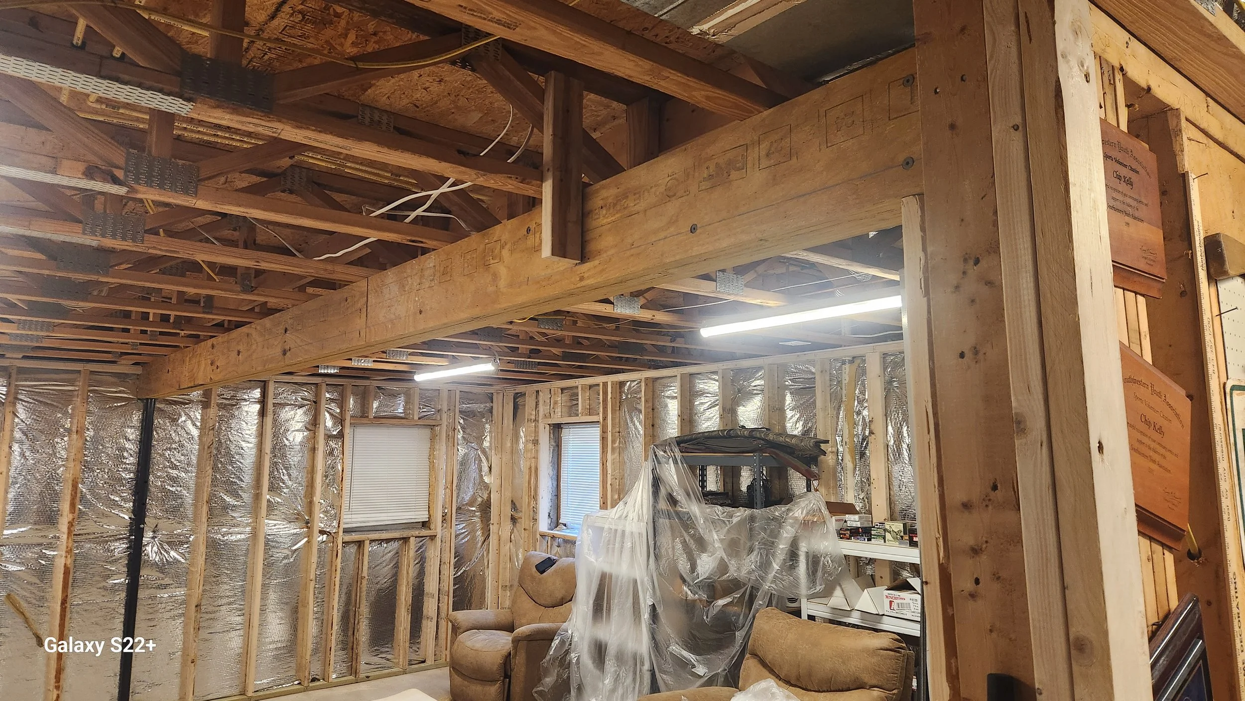 Interior of a house under construction with exposed wooden framing, insulation, and unfinished walls, containing furniture covered in plastic.