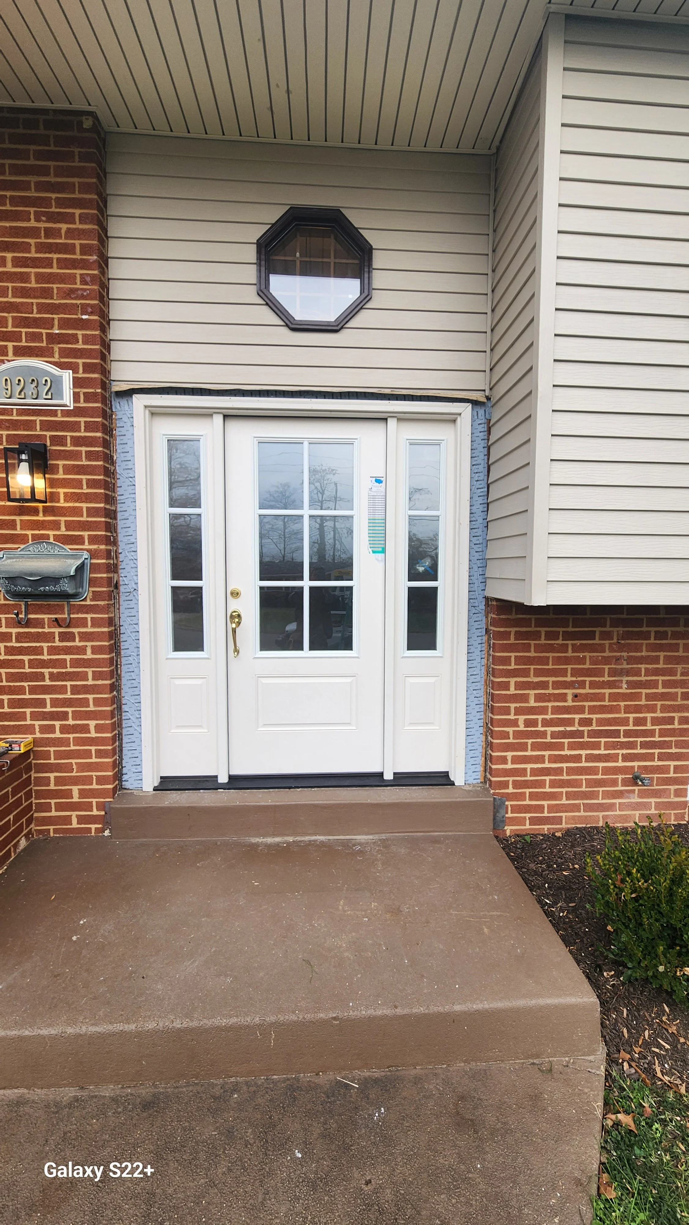 Front entrance of a brick and siding house with a white door with glass panes, a small octagonal window above, and a concrete porch.