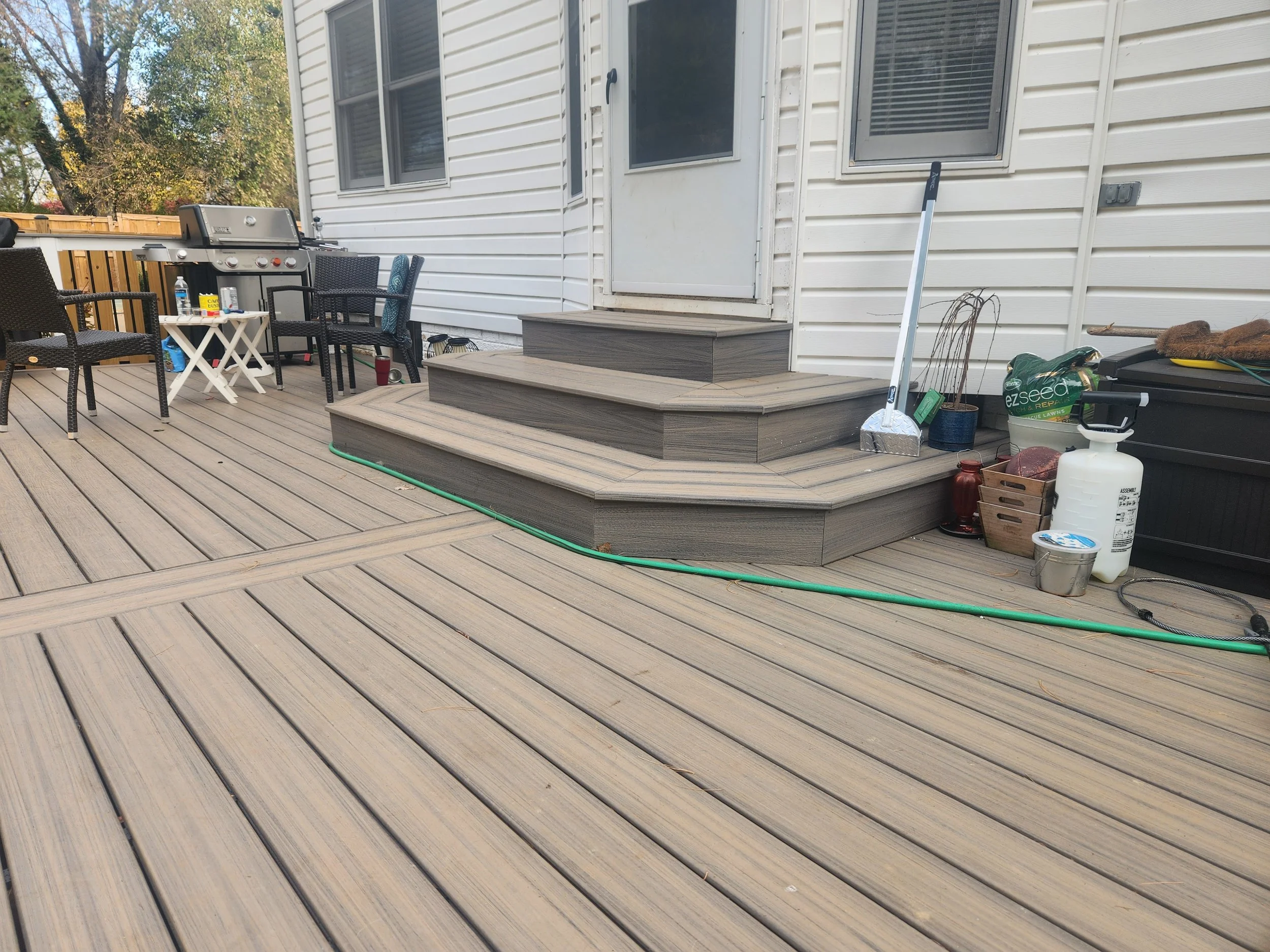 A backyard deck with a set of stairs, outdoor furniture, a grill, and various gardening tools and supplies.