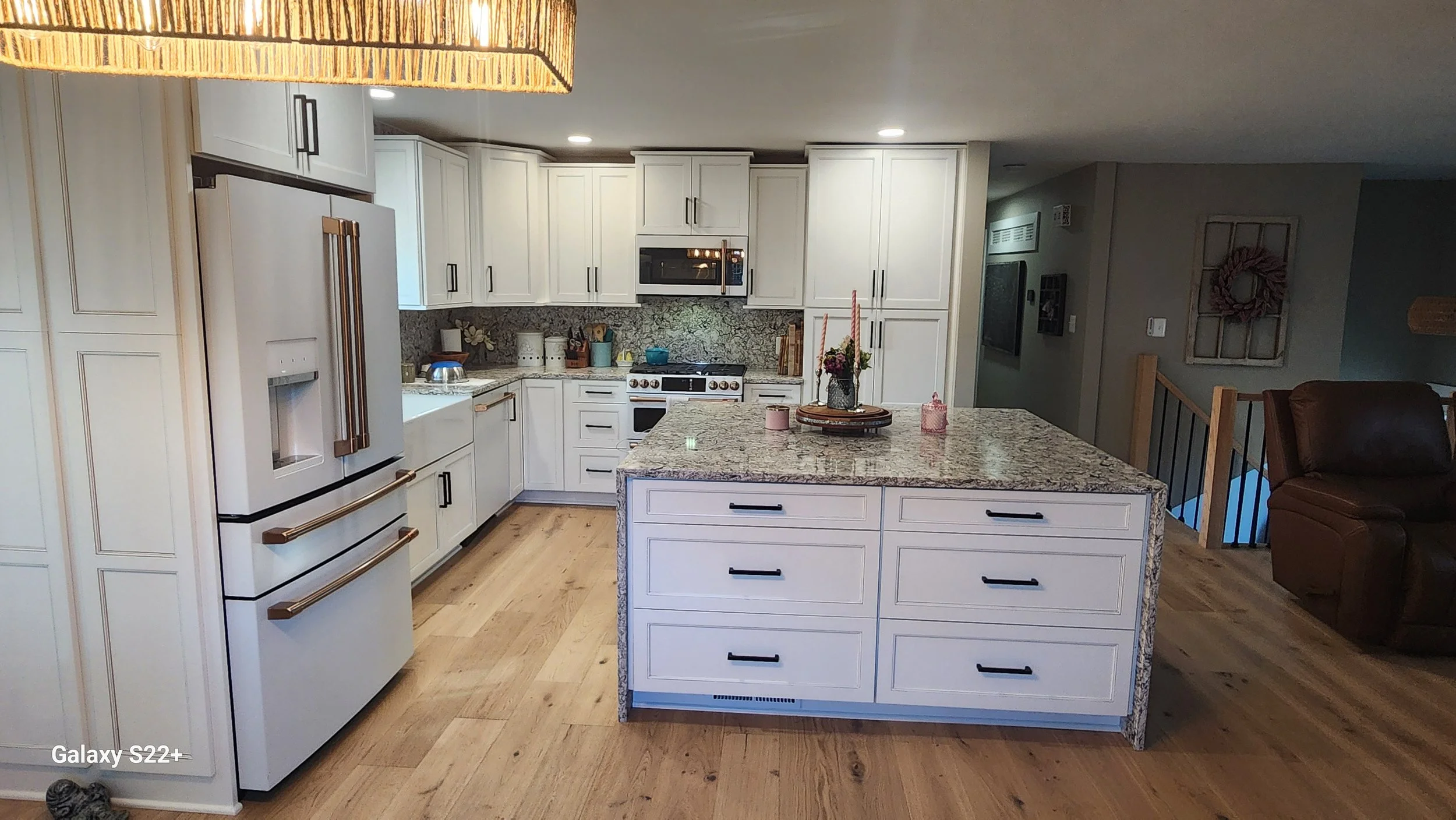 A modern kitchen with white cabinetry, a granite island, stainless steel appliances, wood floors, and a brown leather recliner on the right side.