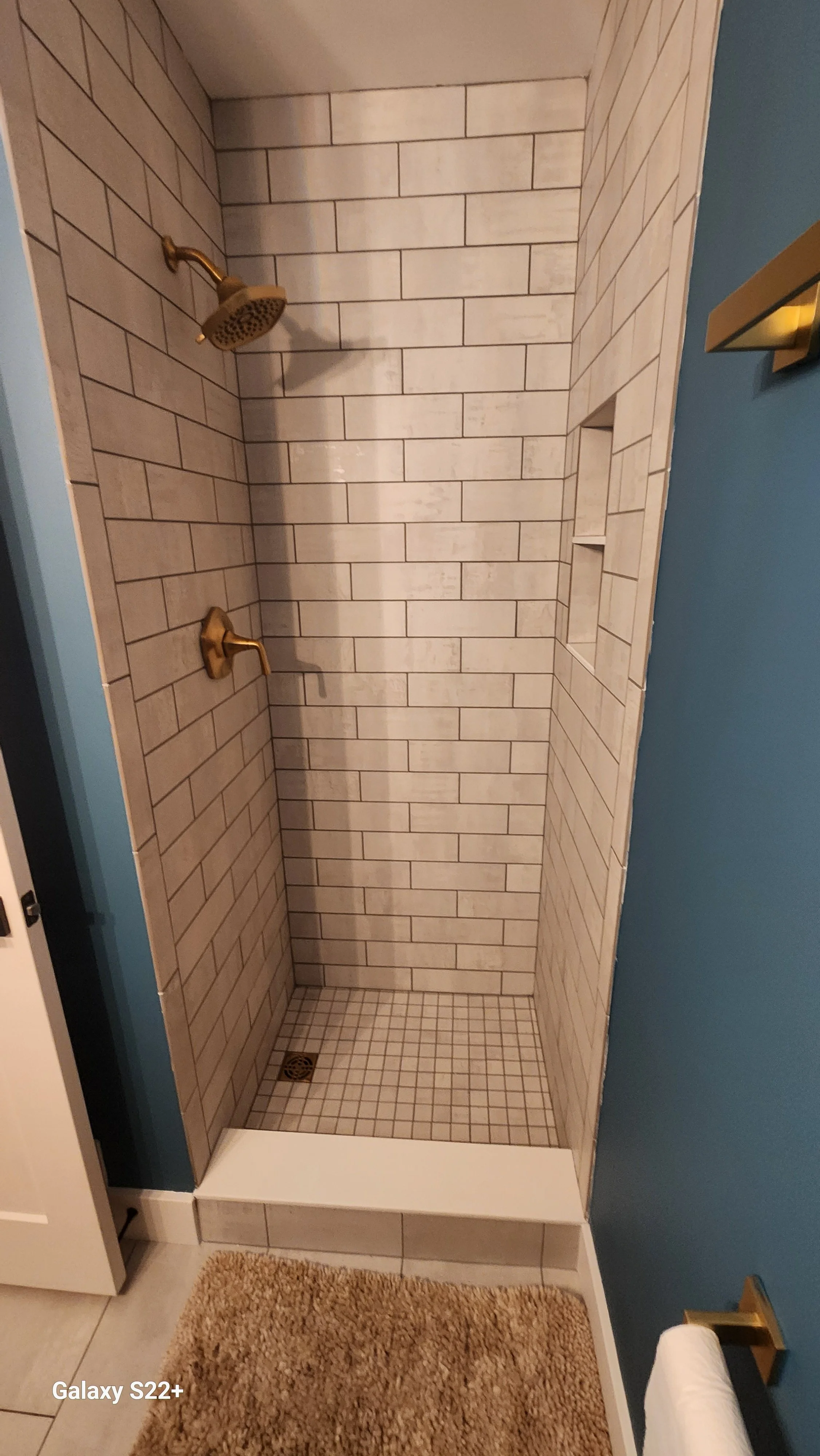 A small, modern shower with beige subway tiles, a brass showerhead and faucet, built-in wall niche, and a beige floor drain, with a blue wall and a towel holder nearby.