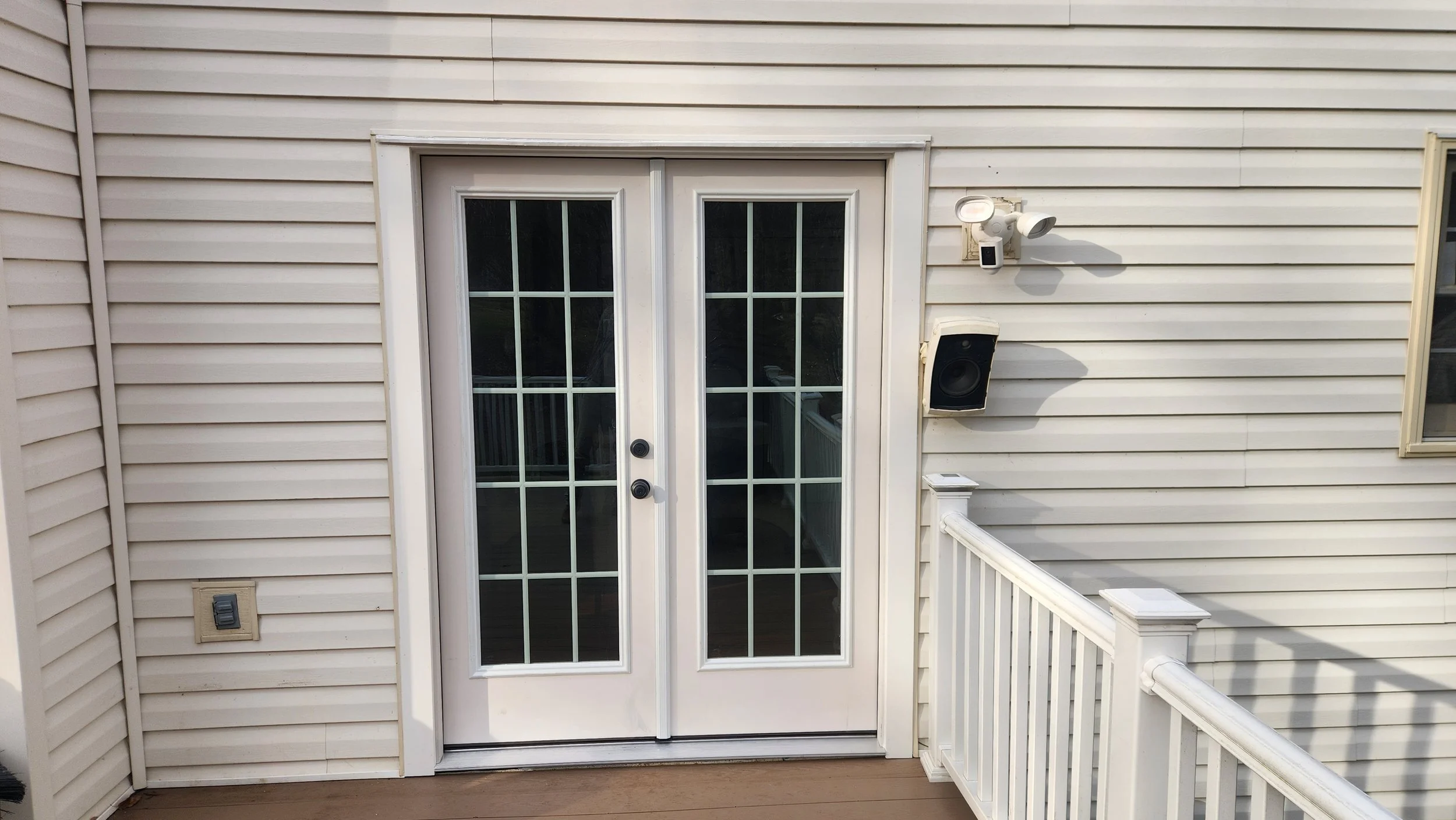Exterior back door with white window-pane double doors, beige siding, wall-mounted security lights, a speaker, and a white railing on a porch.