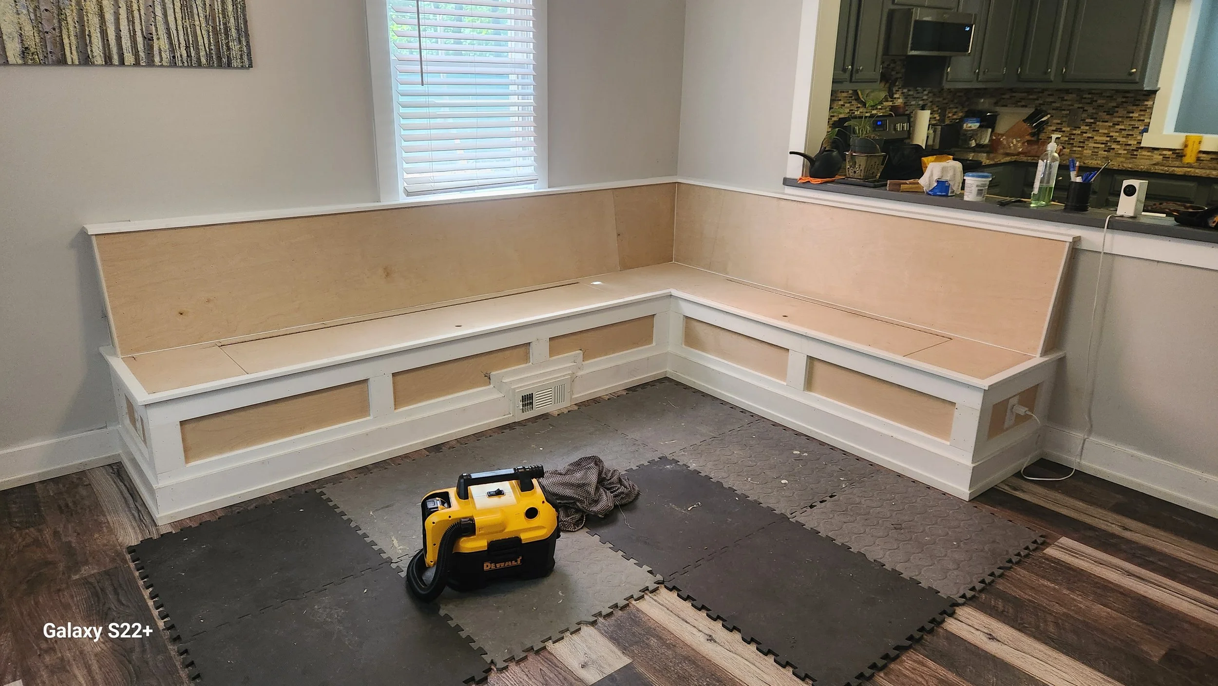 A partially assembled built-in wooden bench with white trim in a kitchen or living room, on a checkered black and gray foam mat, with tools and supplies on the floor and kitchen counter in the background.