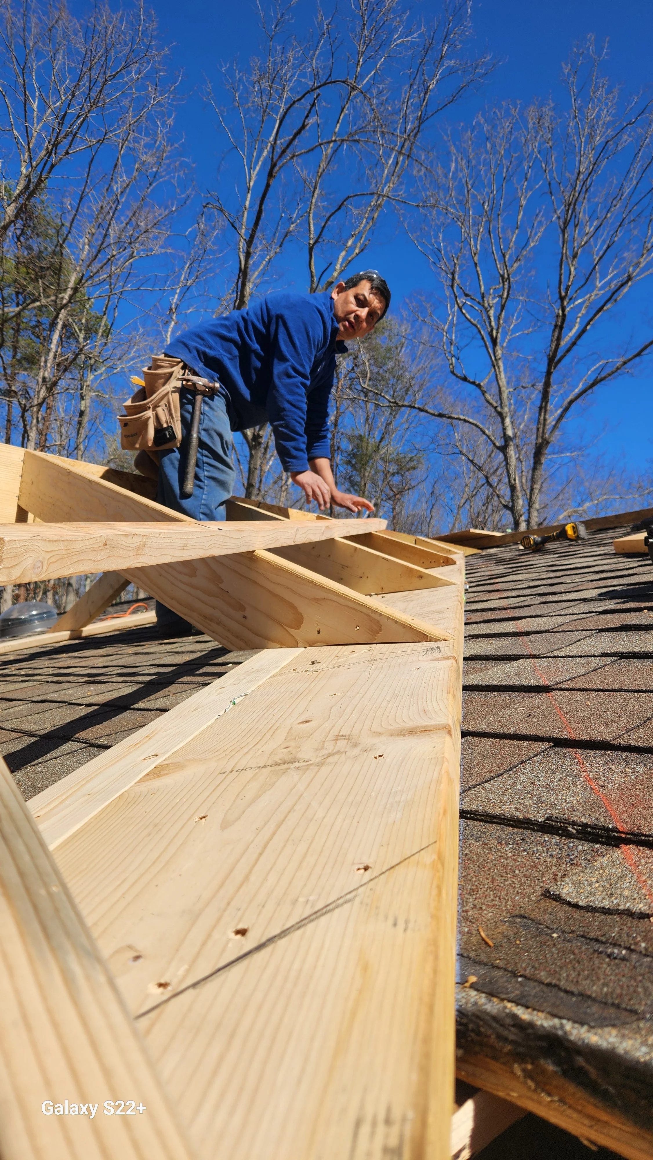 Man working on a roof framing under a clear blue sky, with leafless trees in the background.