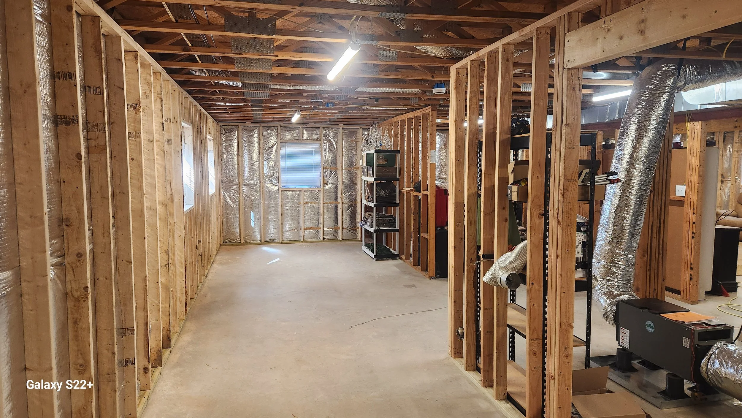 Unfinished basement with wooden framing, insulation, and exposed ceiling beams. There are a few small windows, some shelving units, and ductwork.