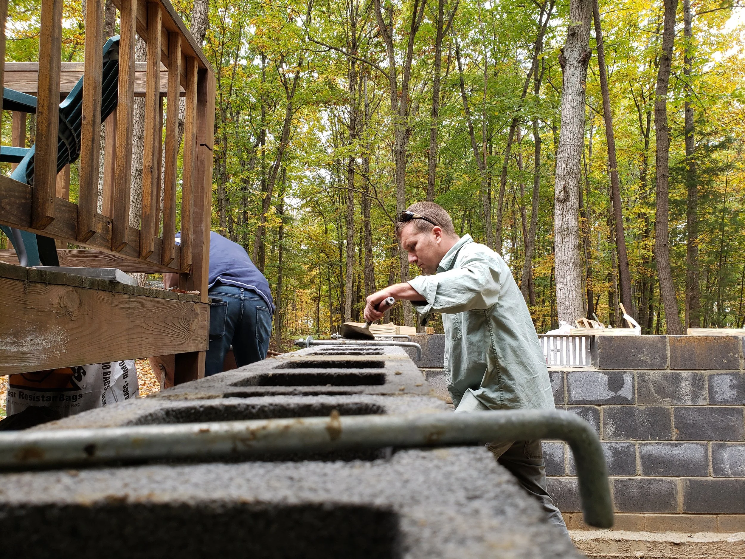 Two men working outdoors in a wooded area with a stone and wooden structure under construction. One is focused on a task, the other is bending down, possibly handling materials.