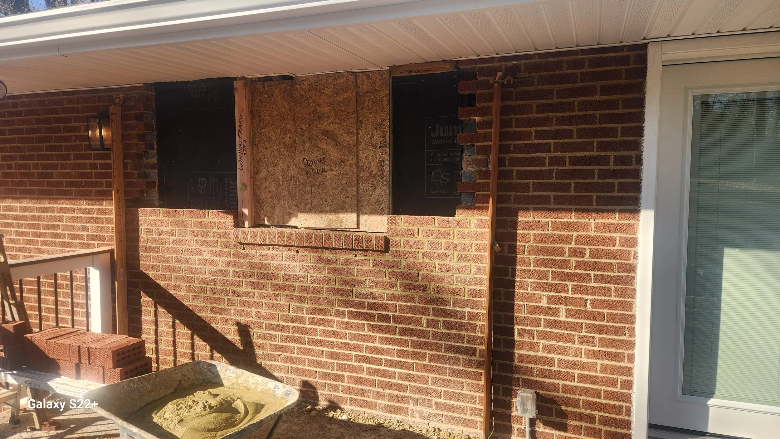 Exterior brick house wall with a door on the right, construction work in progress including plywood covering part of the wall, wooden framing, and a wheelbarrow filled with cement or mortar in the foreground.
