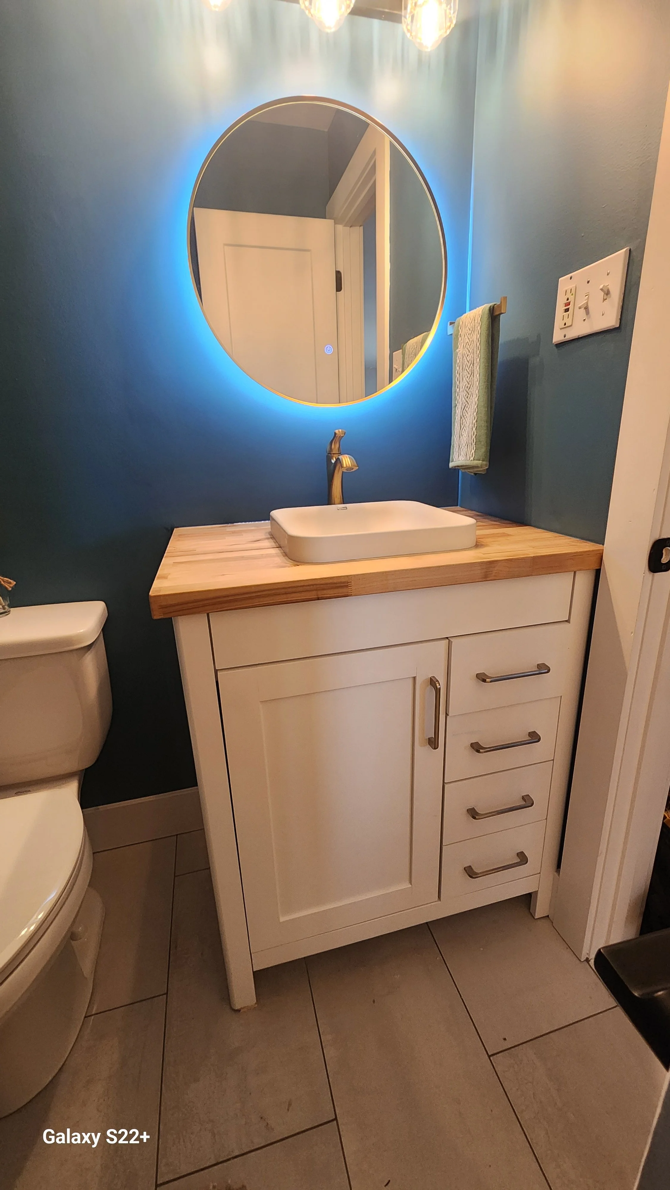 Bathroom with a white cabinet vanity with a wooden countertop, a rectangular vessel sink, a gold faucet, a round mirror with blue backlighting, a towel hanging on a wooden ring, a light switch panel, a toilet to the left, and tiled flooring.