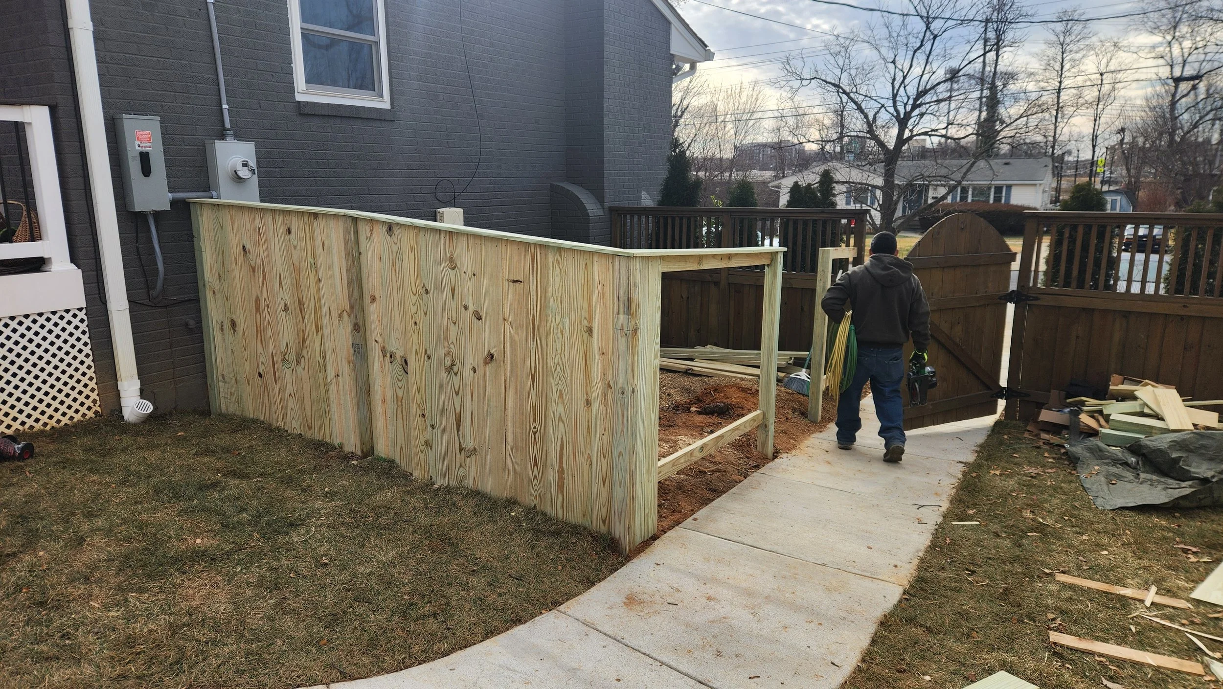 A man is walking on a concrete pathway next to a newly built wooden fence in a backyard. The fence is partially constructed, with some panels and tools visible.