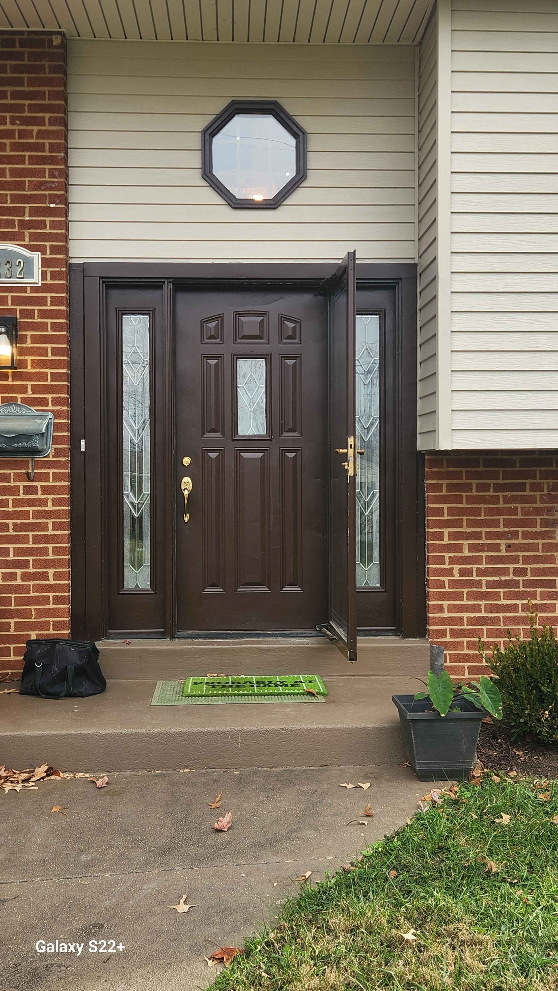 The front porch of a house featuring a black front door with decorative glass panels on either side and above. The house has brick on one side and beige vinyl siding on the other. There is a small green welcome mat, a black bag to the left, and a bla