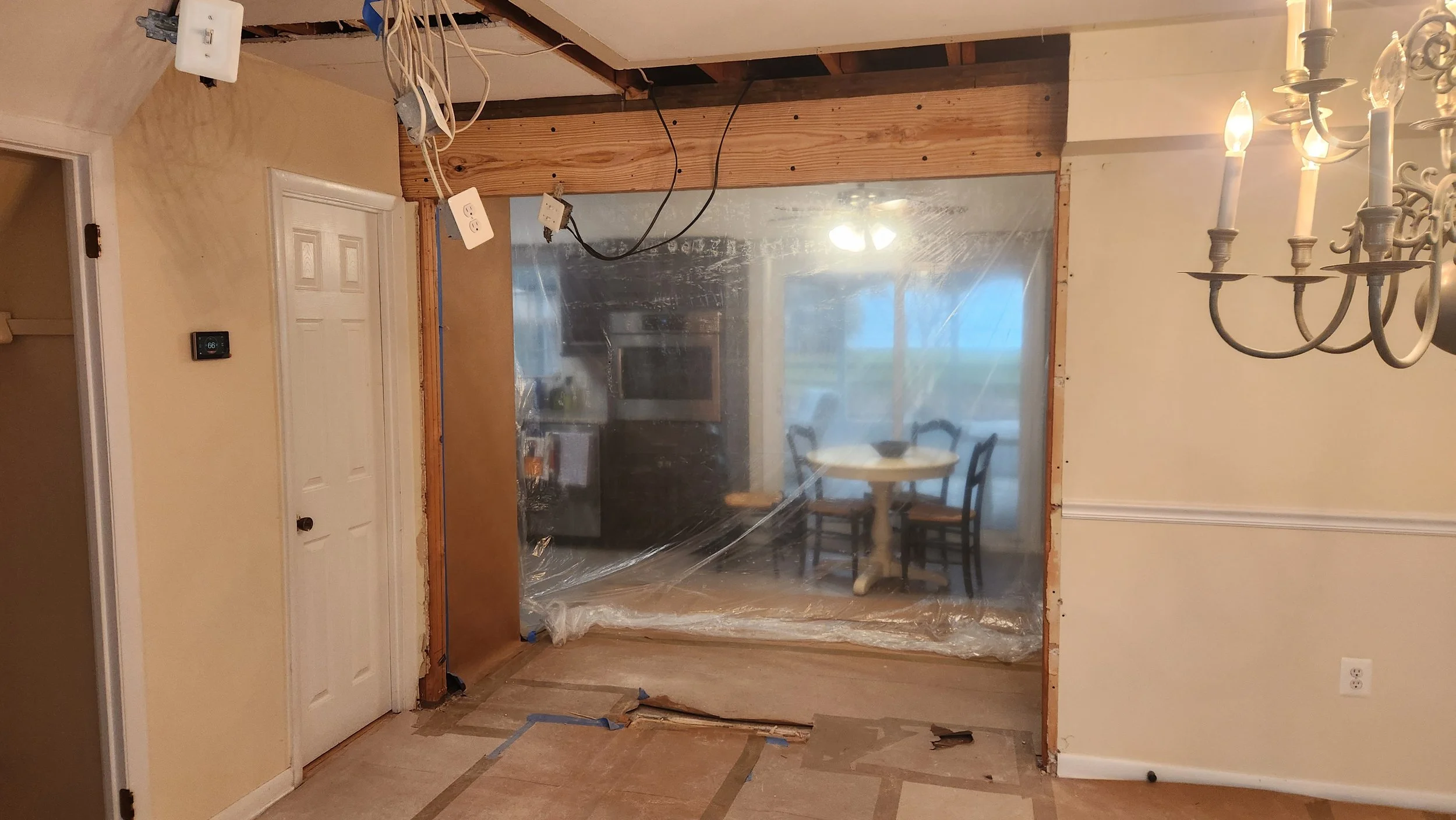 Interior of a kitchen under renovation, with an opening to the dining area covered by a plastic sheet, a chandelier on the right, and electrical wires hanging from the ceiling.