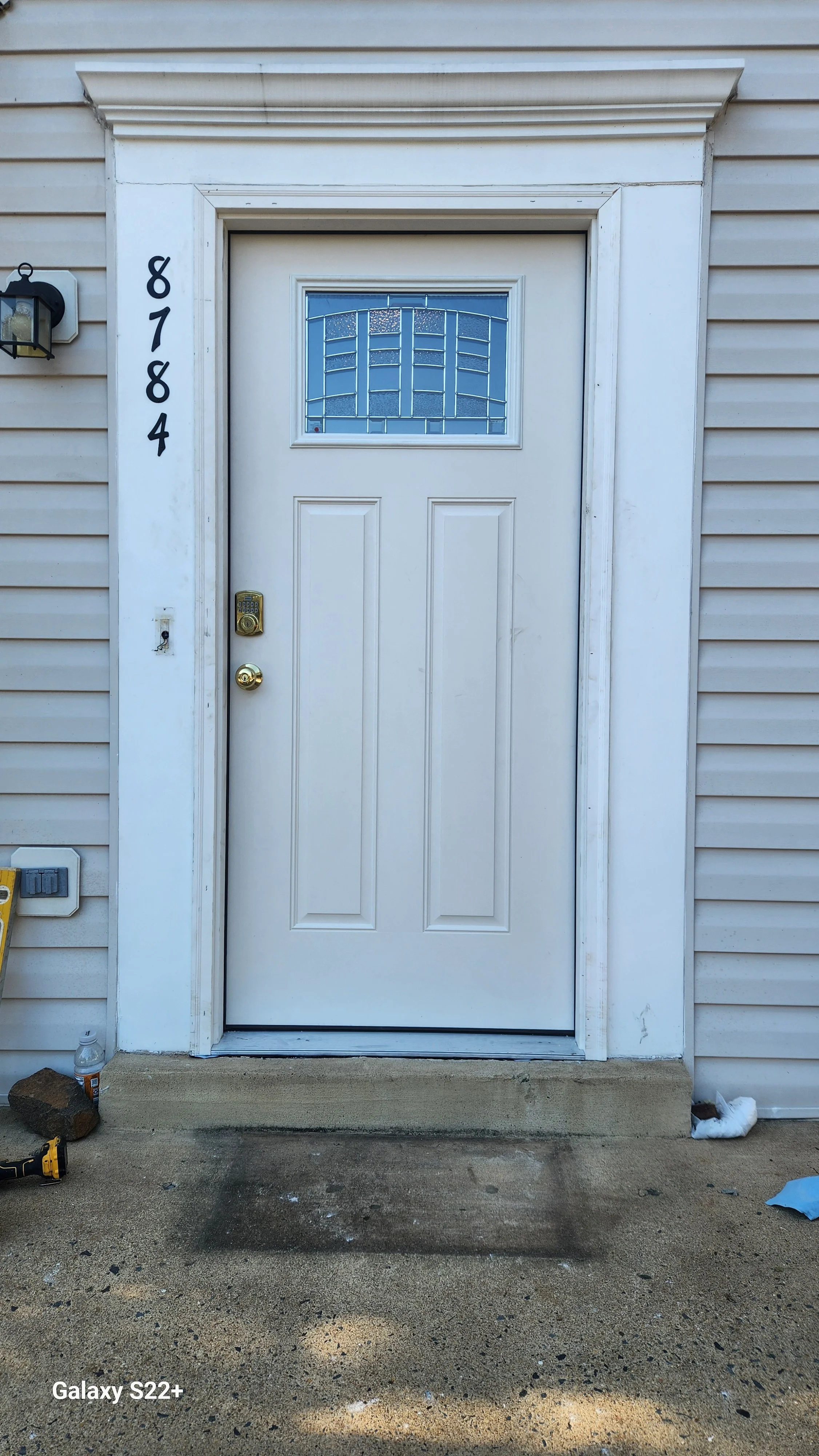 White exterior front door with a glass panel above, beige vinyl siding, house number 8784 on the left, black outdoor light fixture to the left, and construction tools and debris on the concrete porch and ground.