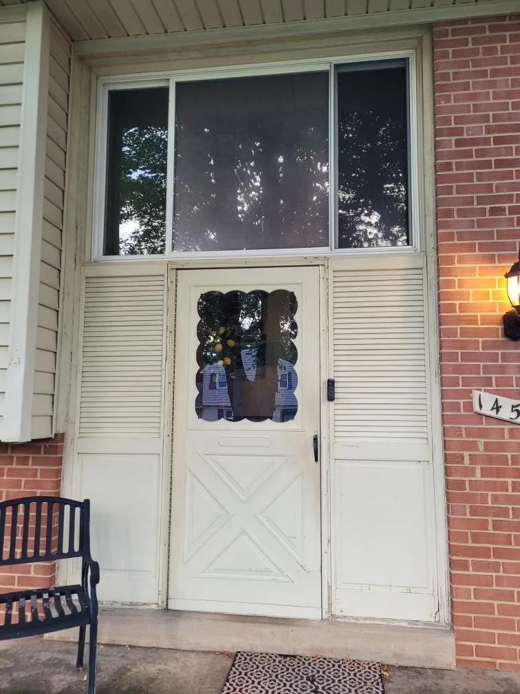 Front porch with a white door featuring a decorative cutout, surrounded by beige and brick siding. A black bench is on the left, a small patterned doormat is in front, and a wall-mounted lamp with a warm glow is on the right.