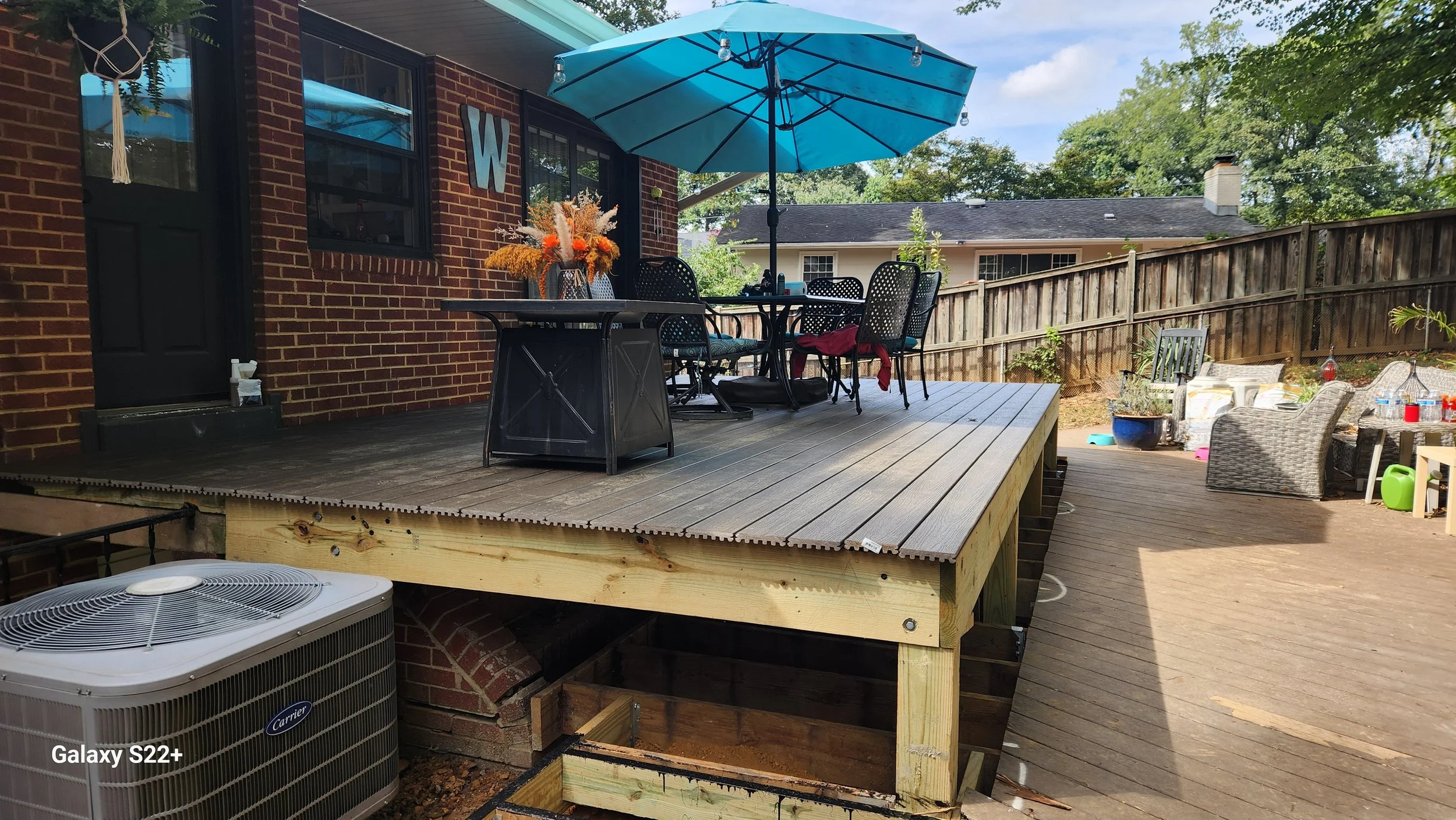 Backyard patio with a raised wooden deck, outdoor dining table with black chairs, a large blue umbrella, and other outdoor furniture, with a brick house wall and a wooden fence in the background.